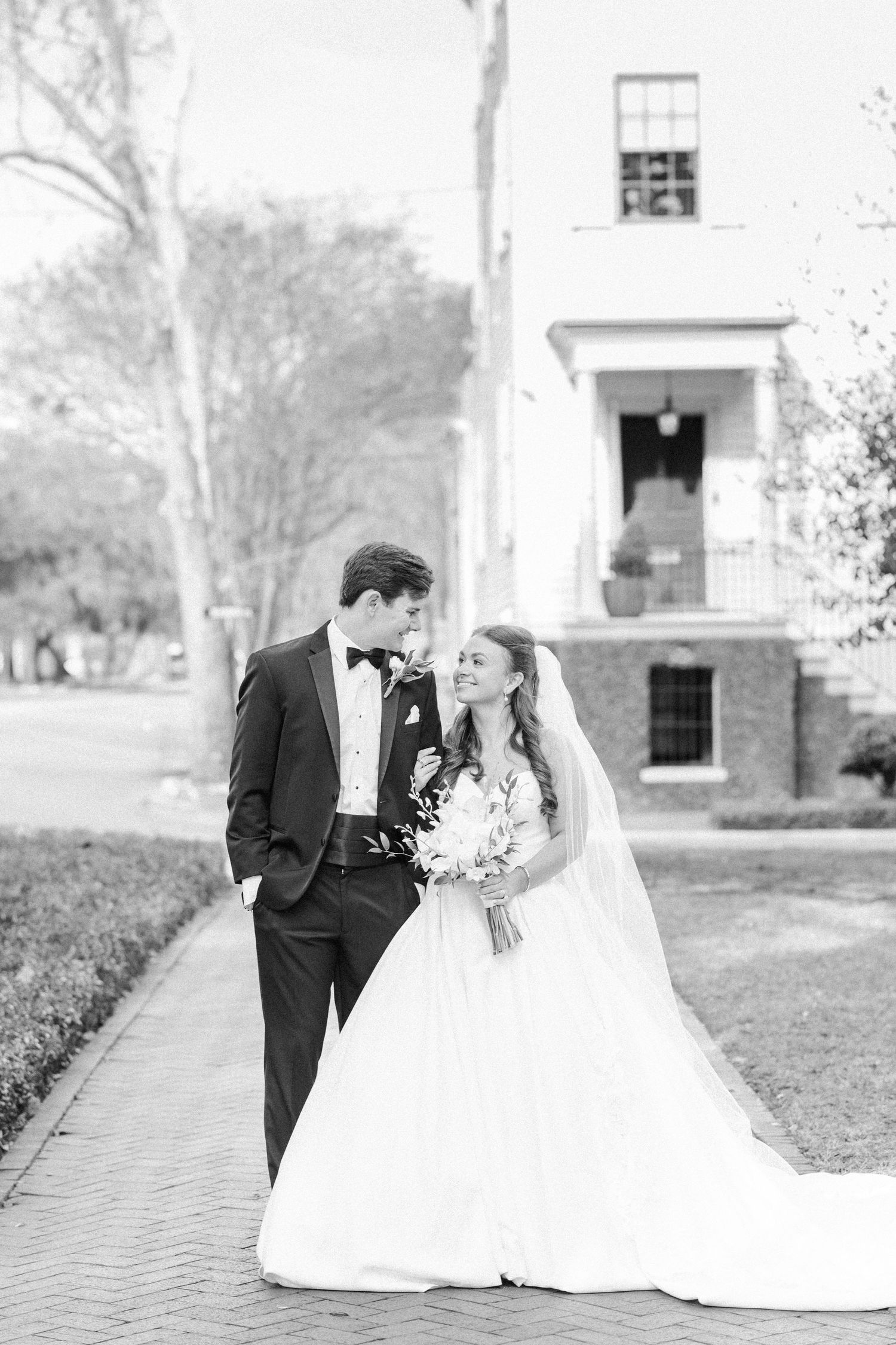 A black and white wedding photo of a couple sharing a romantic moment on a brick path outside a classic building.