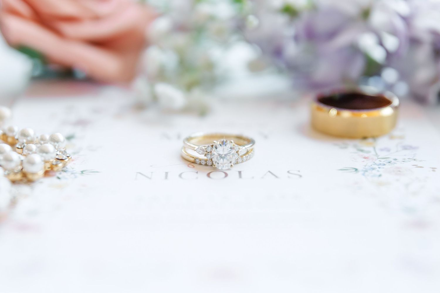Wedding rings and jewelry styled on white surface with soft pink and purple floral accents.