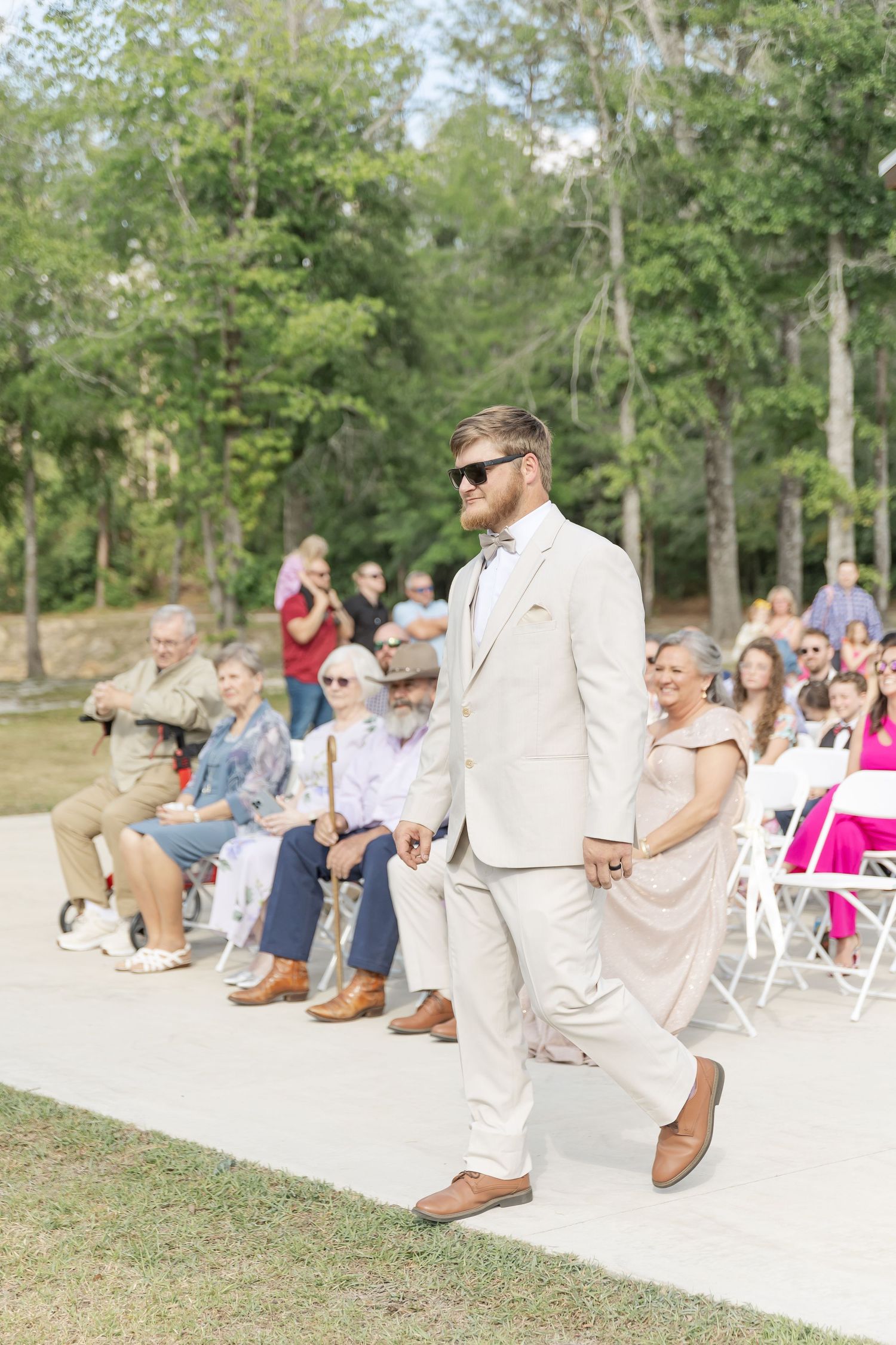 Wedding attendee in a beige suit walks down an outdoor aisle with guests seated on white chairs.