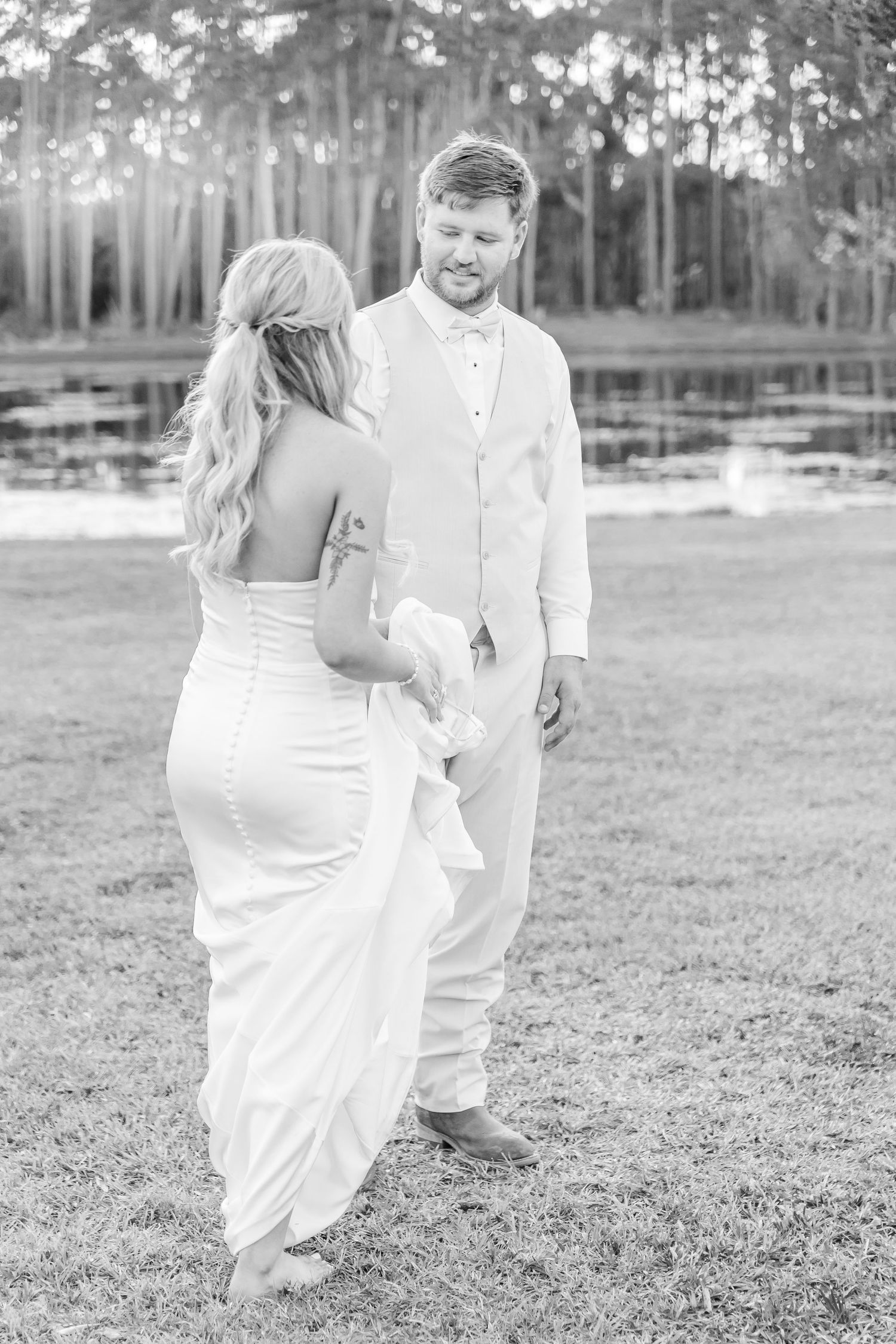 Black and white wedding photo of couple in elegant white attire standing by a lake.