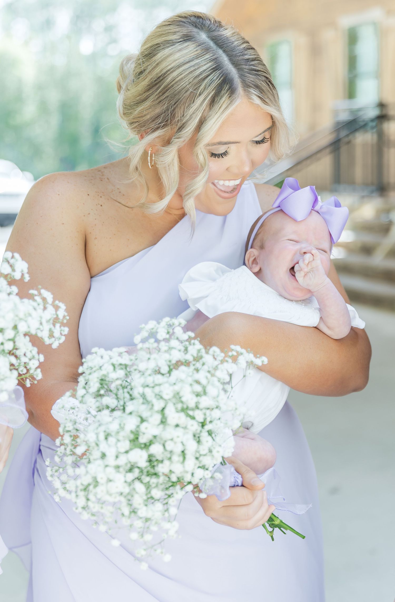 Bridesmaid in lavender dress holds baby while carrying white floral bouquet.