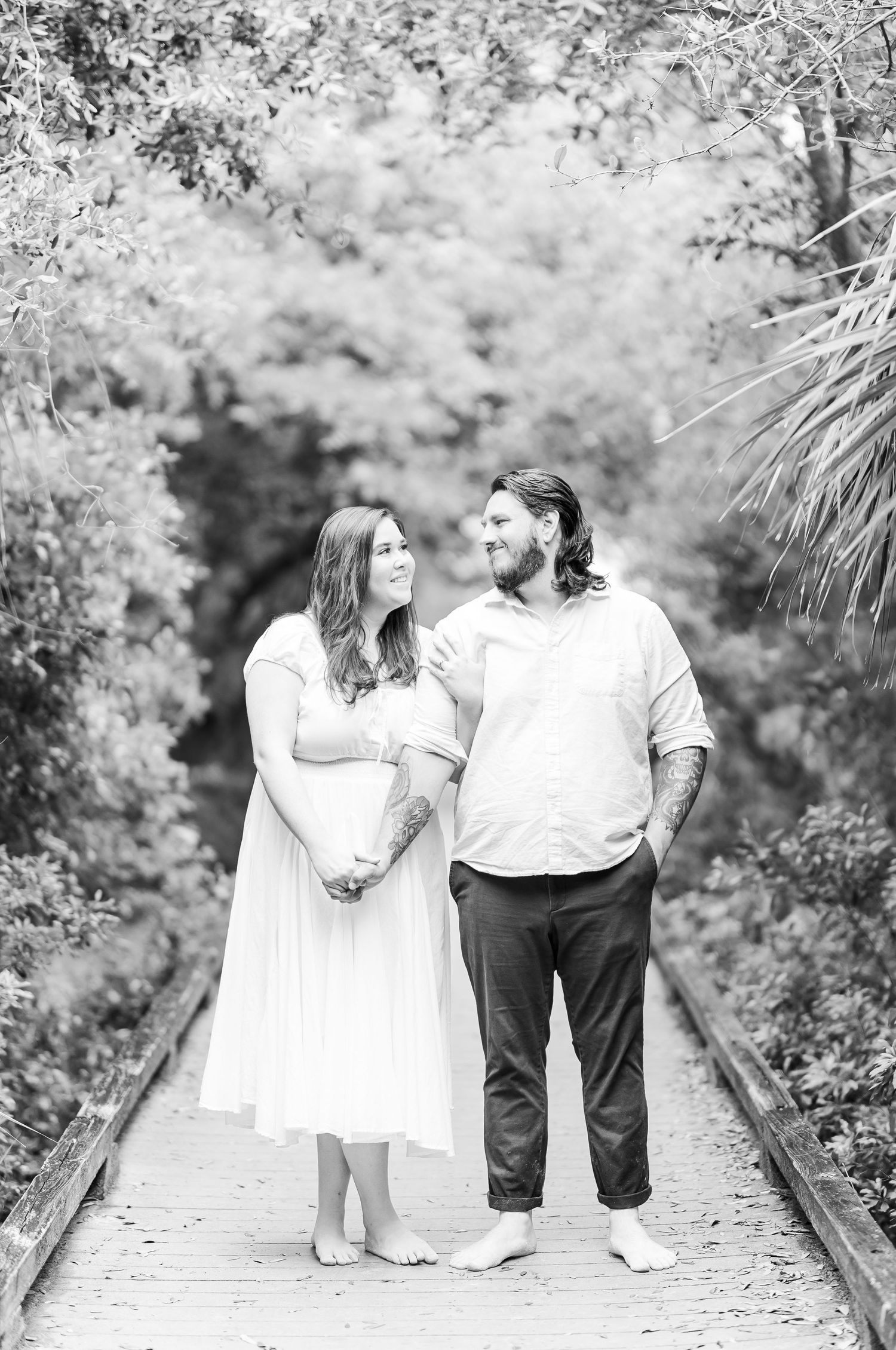 A couple stands together on a wooden boardwalk surrounded by trees in black and white photography.