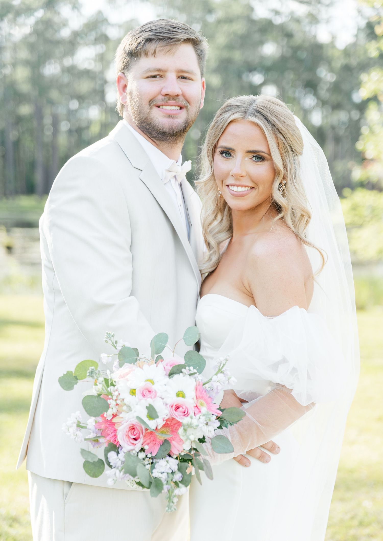 A joyful wedding portrait of a couple in white attire against a natural outdoor backdrop.