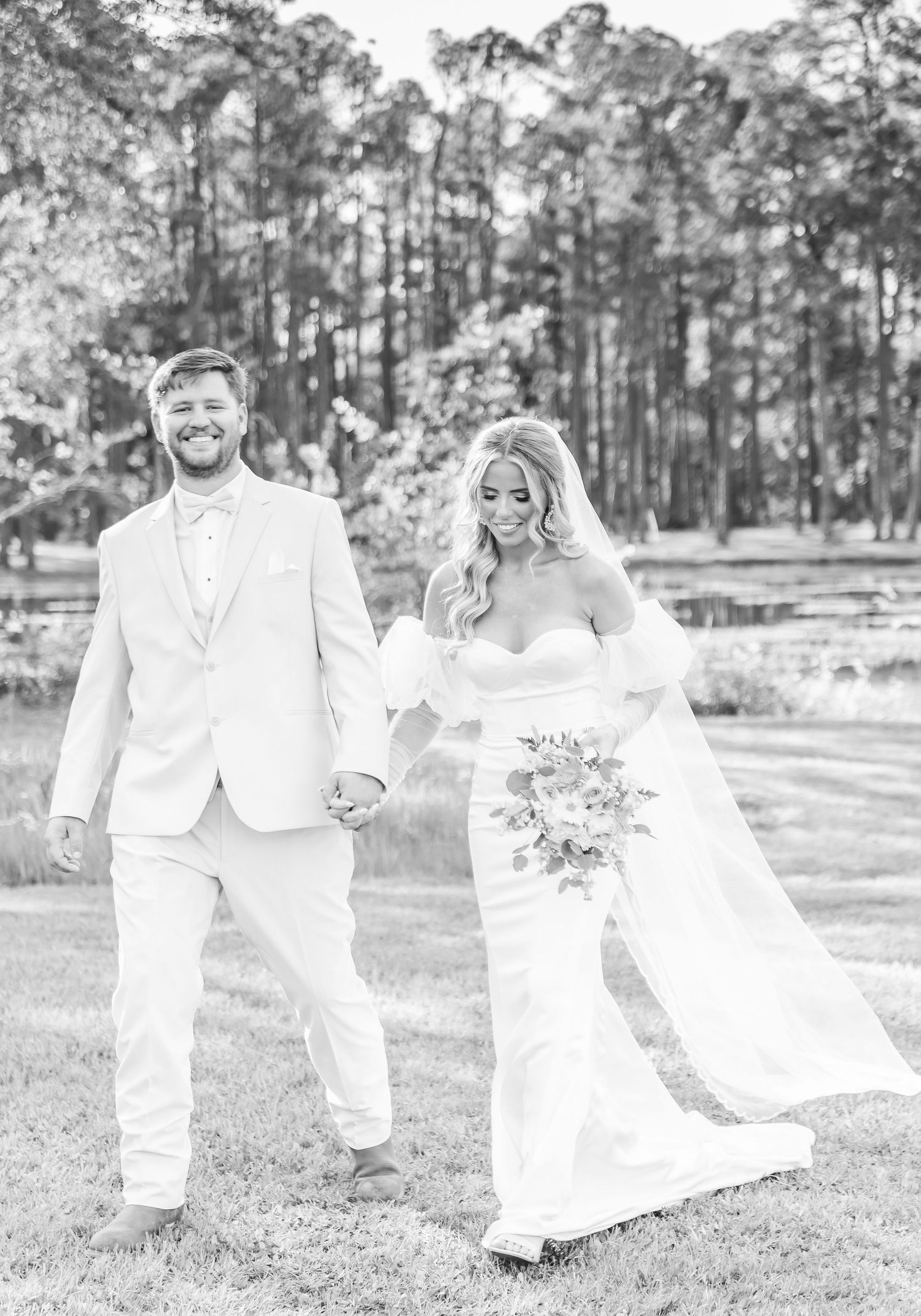 Black and white portrait of a wedding couple walking together in a pine forest setting.