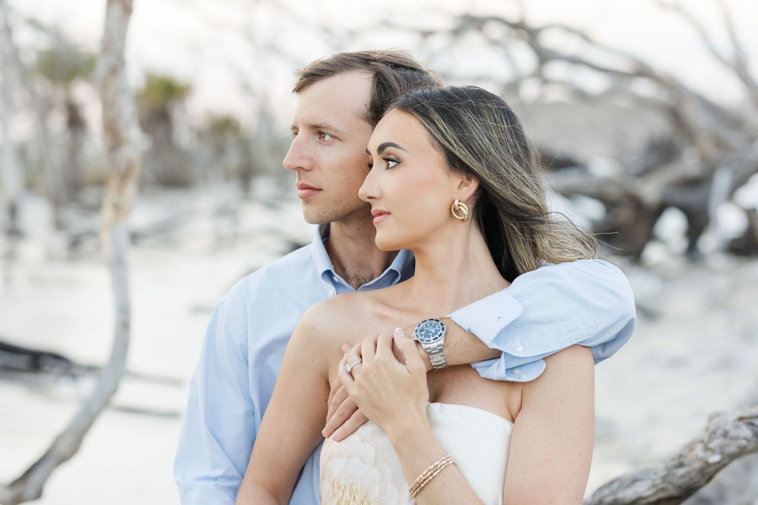 Couple embracing near driftwood on beach, woman in white strapless top wearing blue watch.
