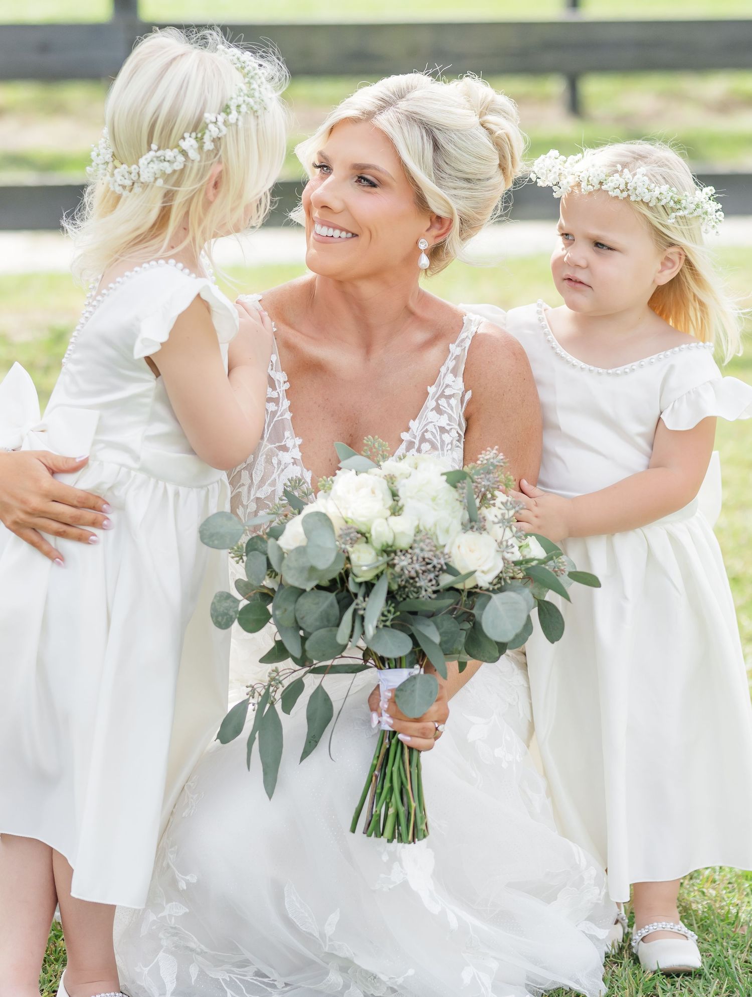 A bride in a lace wedding dress holds a eucalyptus bouquet while kneeling with two flower girls in white dresses and floral crowns.
