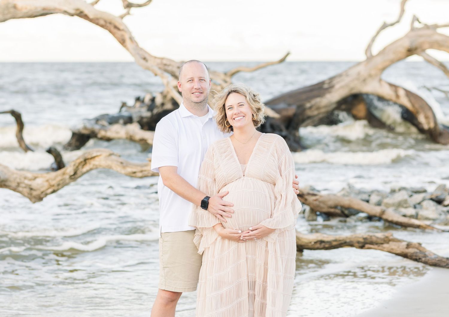 A couple wearing white and blush outfits embraces lovingly on a beach with driftwood and waves in the background.