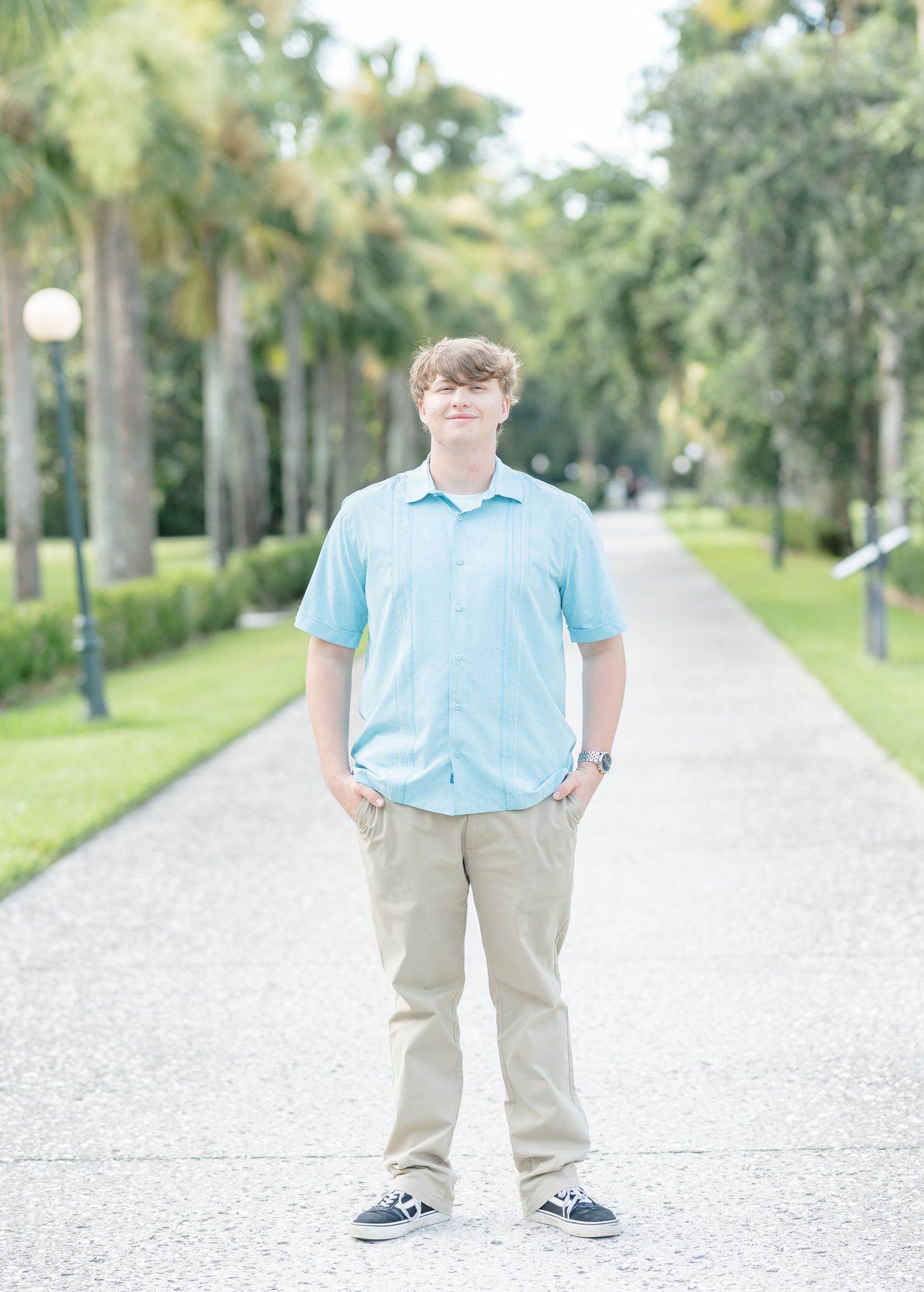 Person wearing a light blue shirt and khaki pants stands on a pathway lined with palm trees.