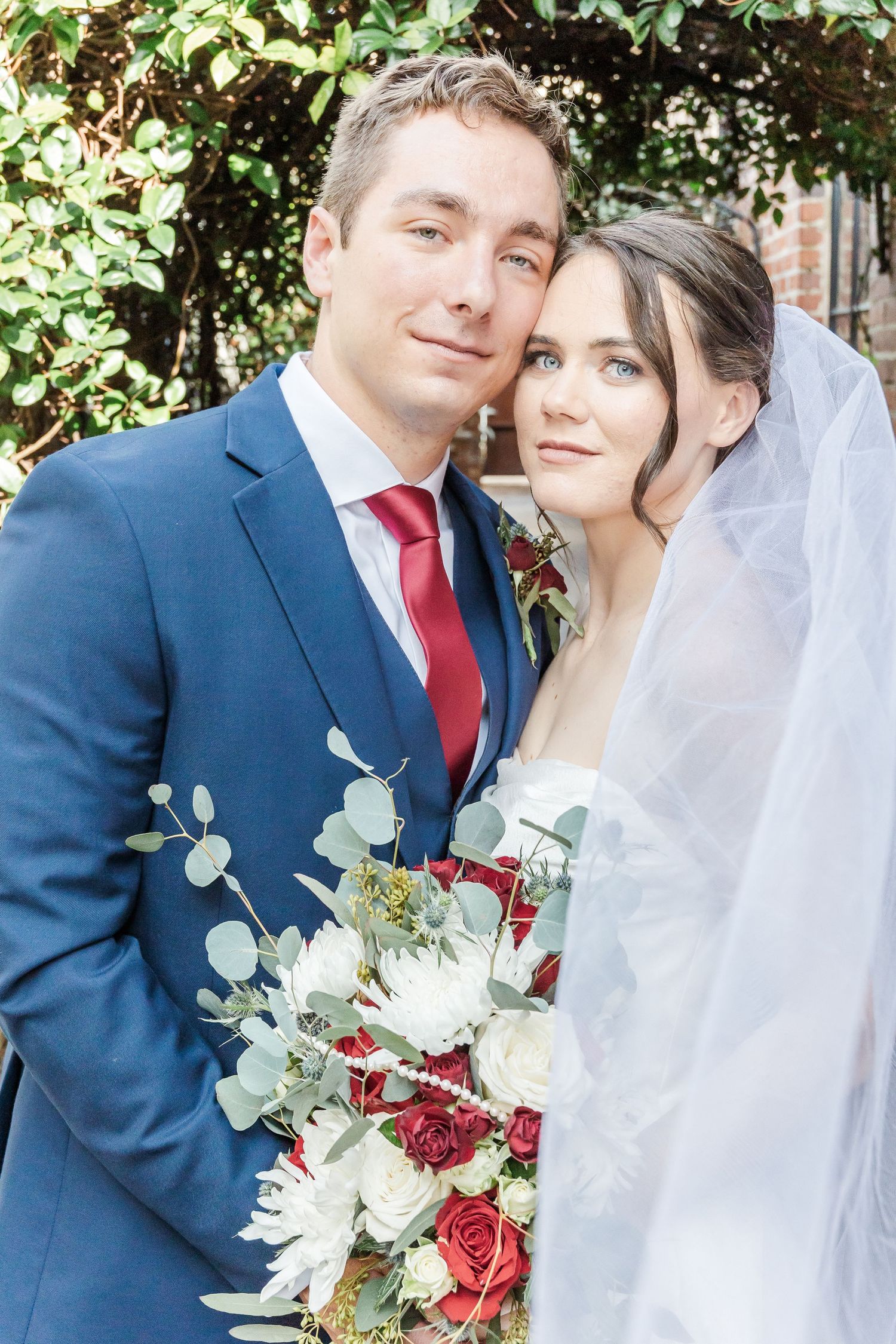 A wedding couple embraces outdoors wearing navy suit and white gown with red and white floral bouquet.