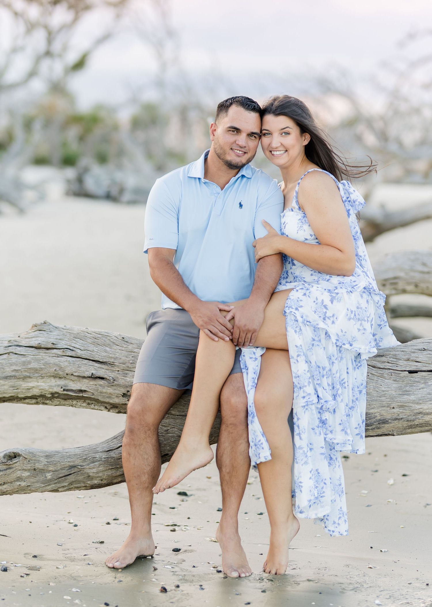 A couple poses together on a beach in coordinating outfits - a blue polo with grey shorts and a floral blue dress.