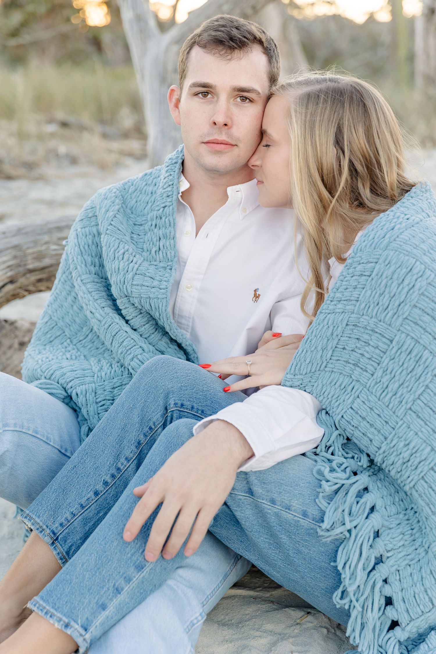 A couple in matching blue sweaters and jeans sits close together on the beach during an intimate photo session.