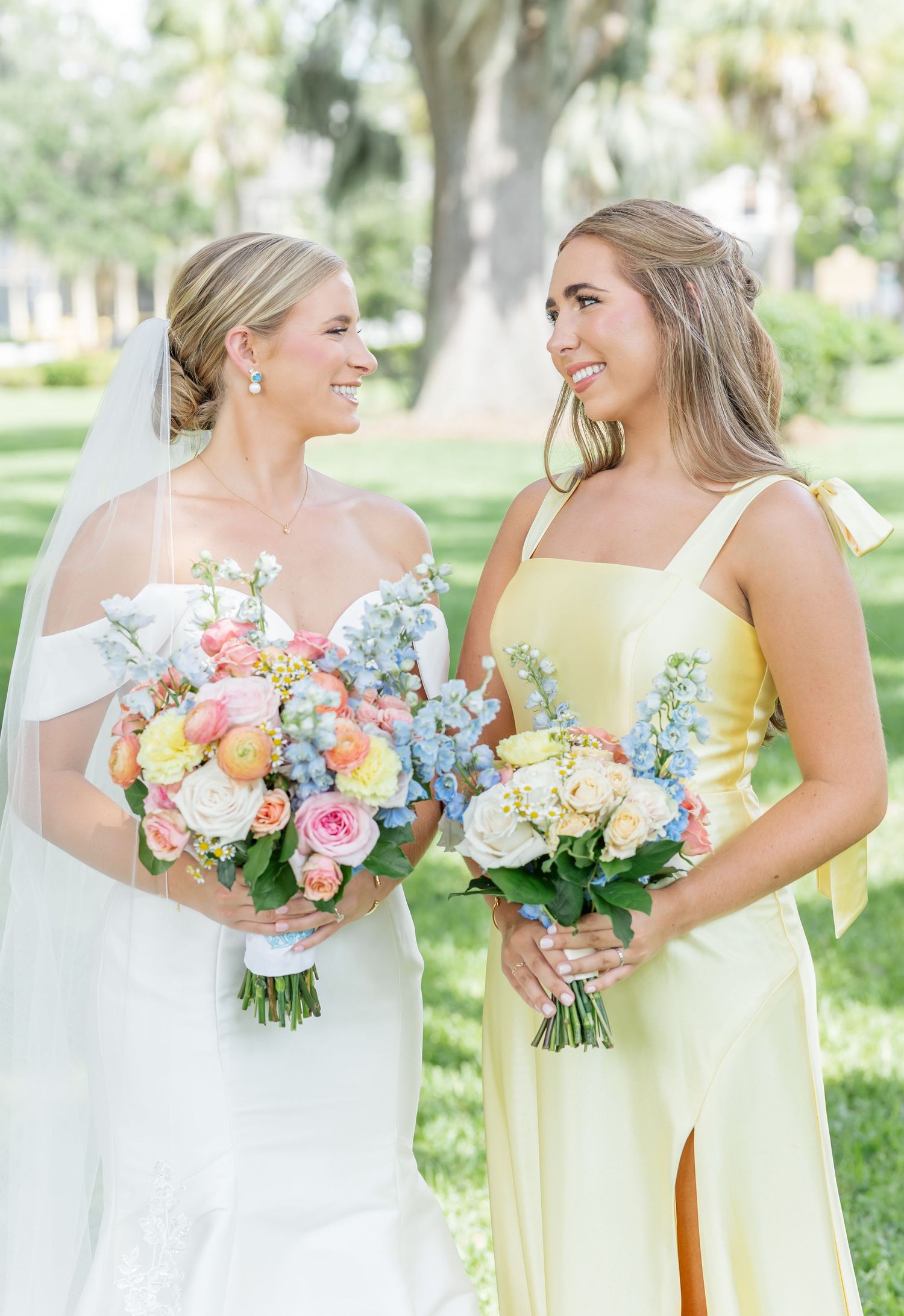 Two wedding attendants hold colorful floral bouquets while sharing a joyful moment outdoors.