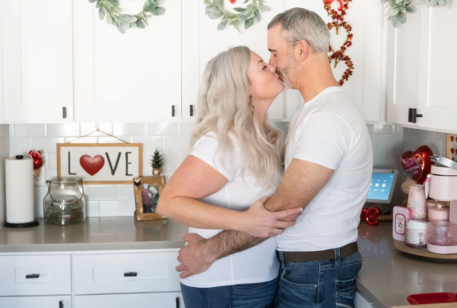 A couple embraces in their kitchen with holiday decorations and a 'LOVE' sign visible in the background.