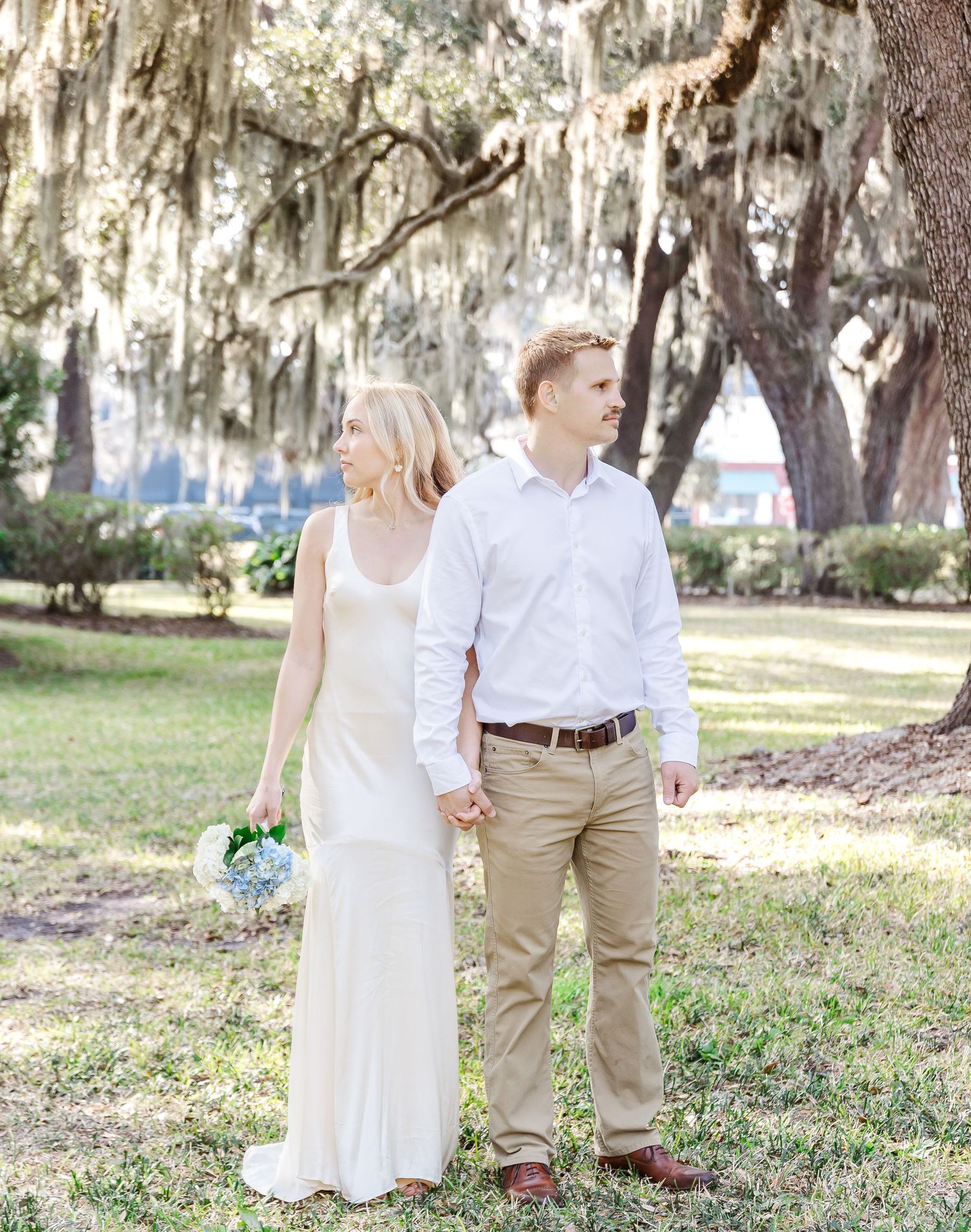 A couple strolls hand in hand through a garden with Spanish moss hanging overhead.