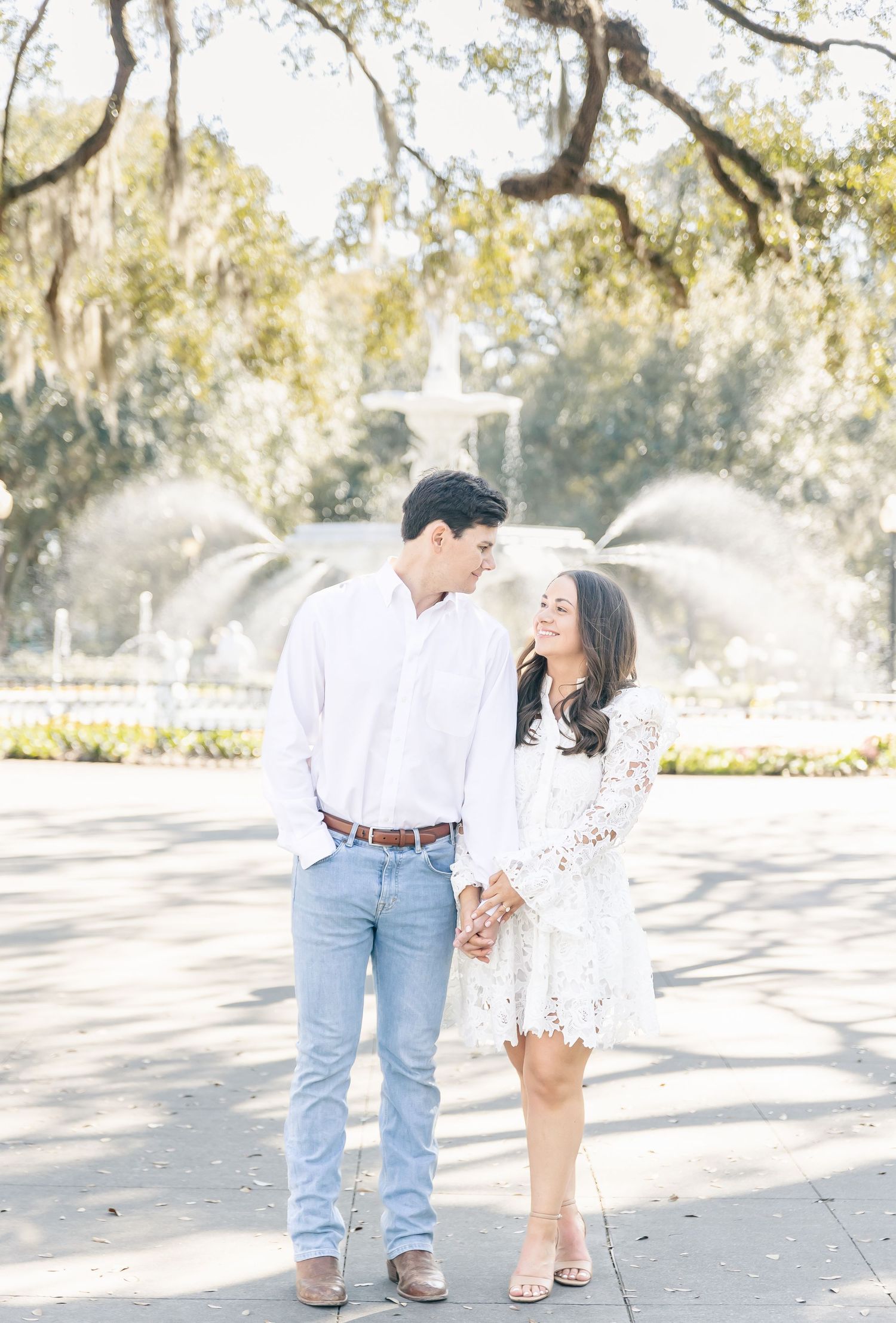 A romantic couple poses for engagement photos in front of a garden fountain with moss-draped trees.