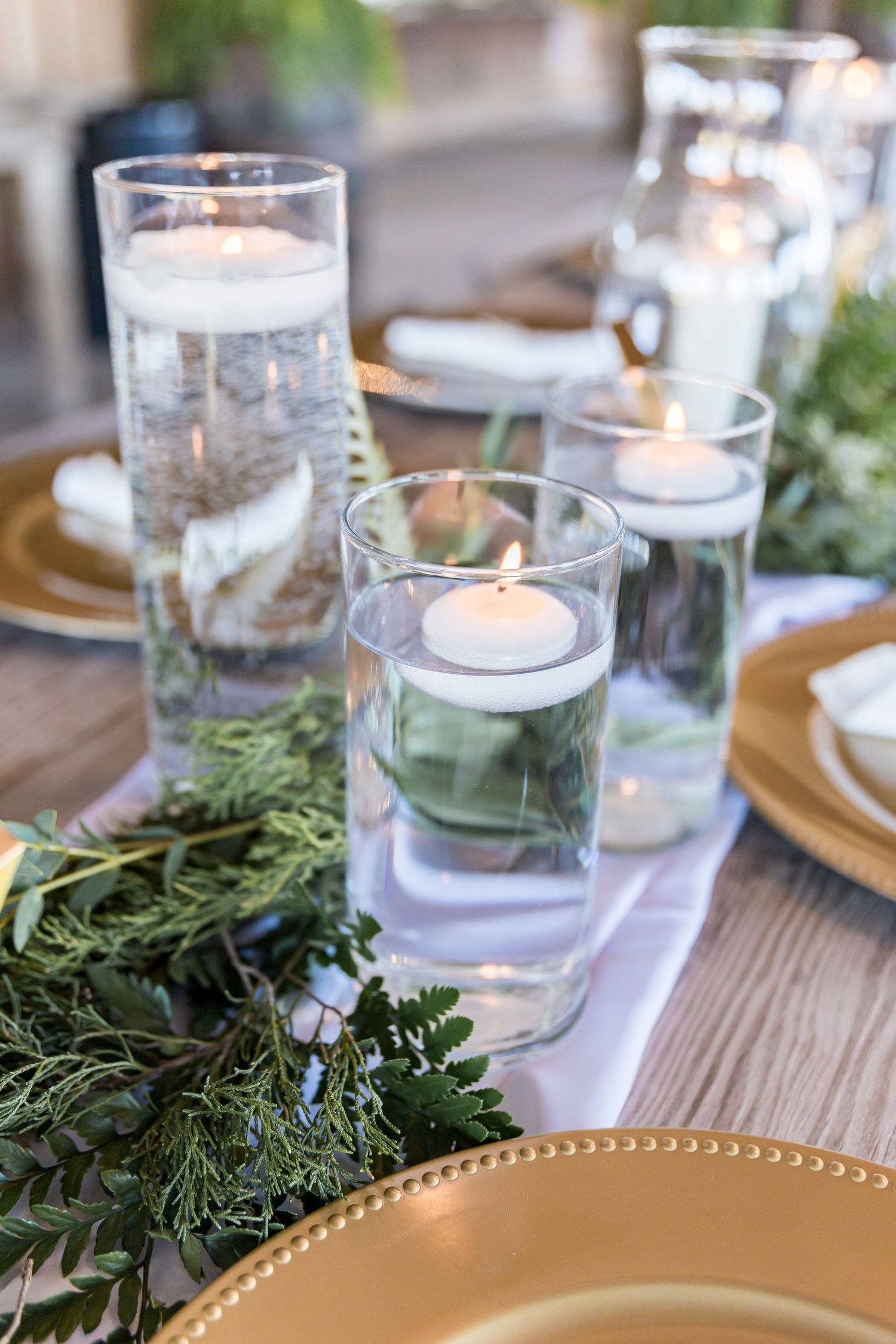 Floating candles in glass cylinders with greenery and gold charger plates on wooden table.