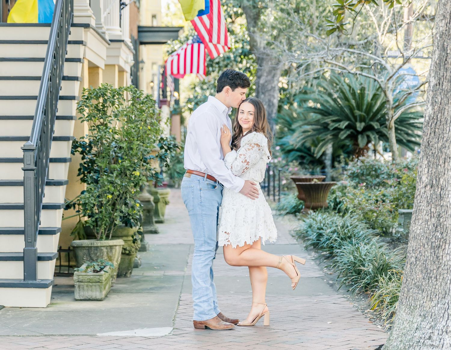 Sweet engagement moment captured on brick sidewalk with woman in white lace dress.