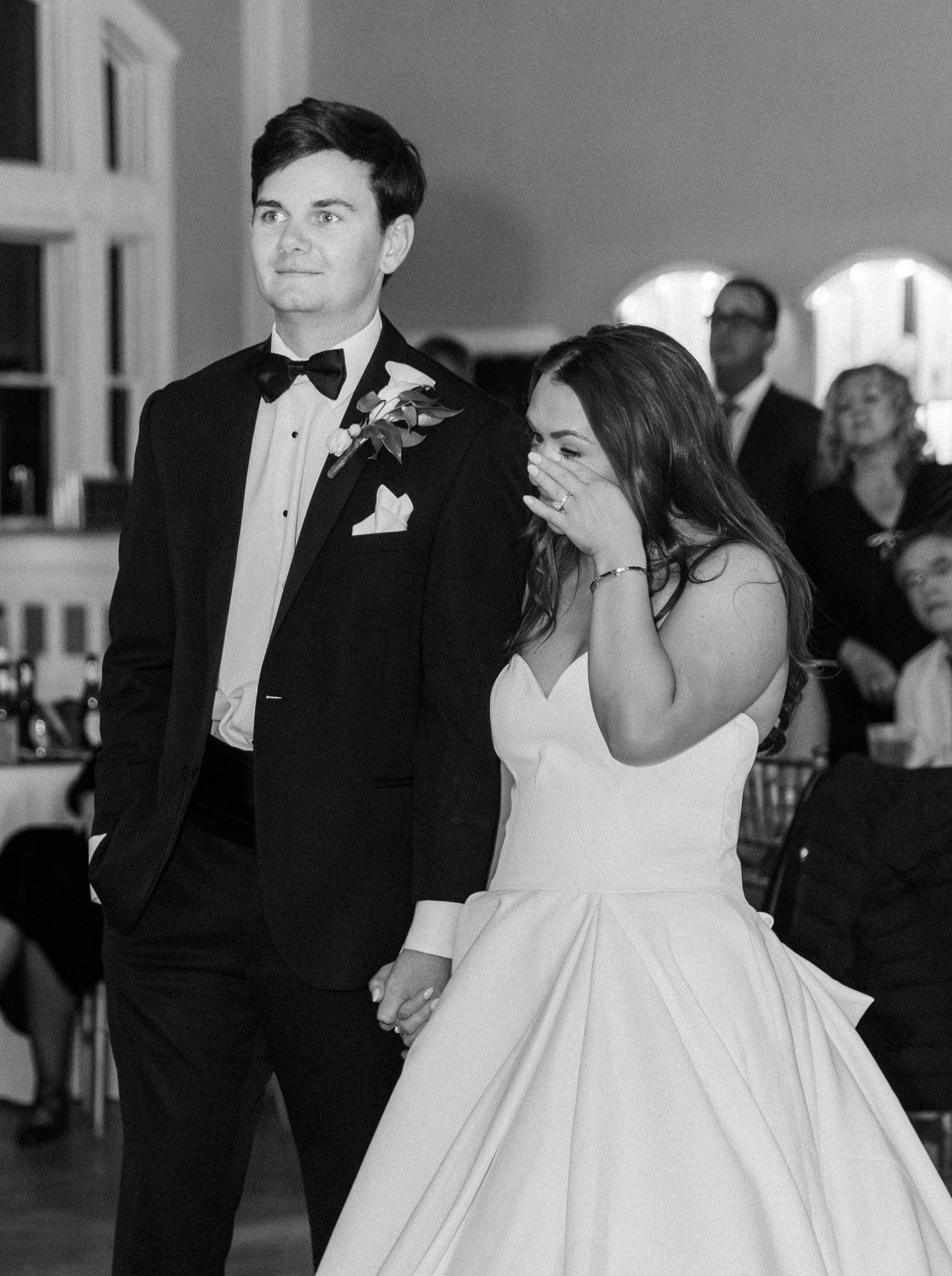 Black and white photo of a bride wiping tears while holding hands with her groom in a tuxedo at their wedding reception.