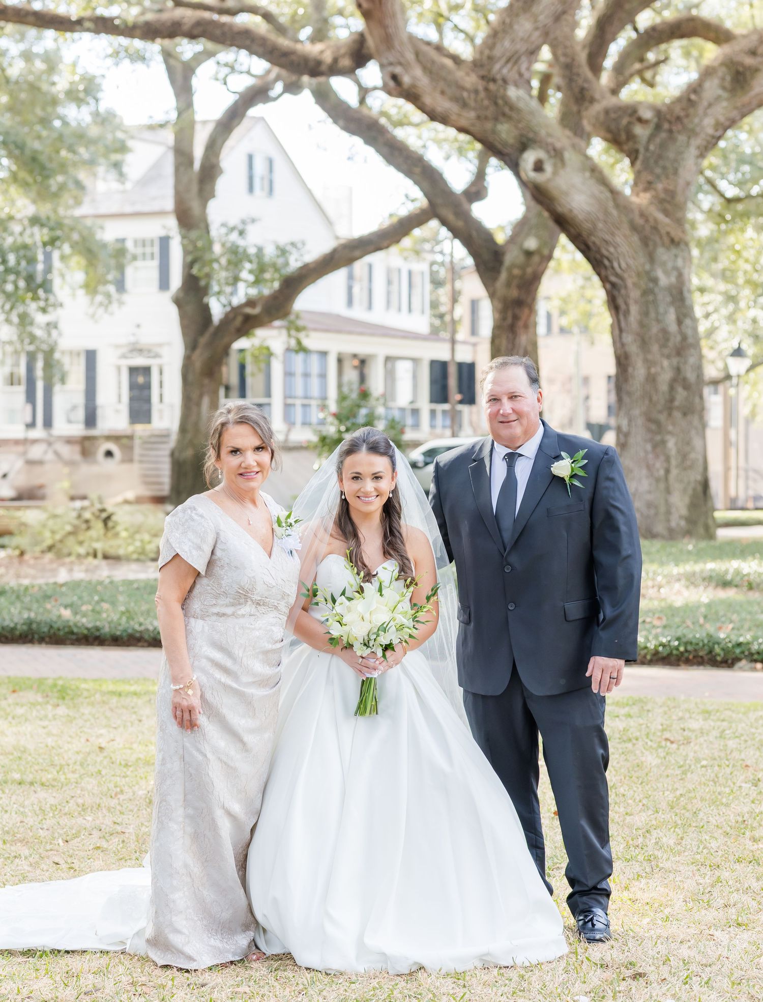 Bride in white ball gown with veil poses with parents in formal attire beneath large oak trees at outdoor wedding.