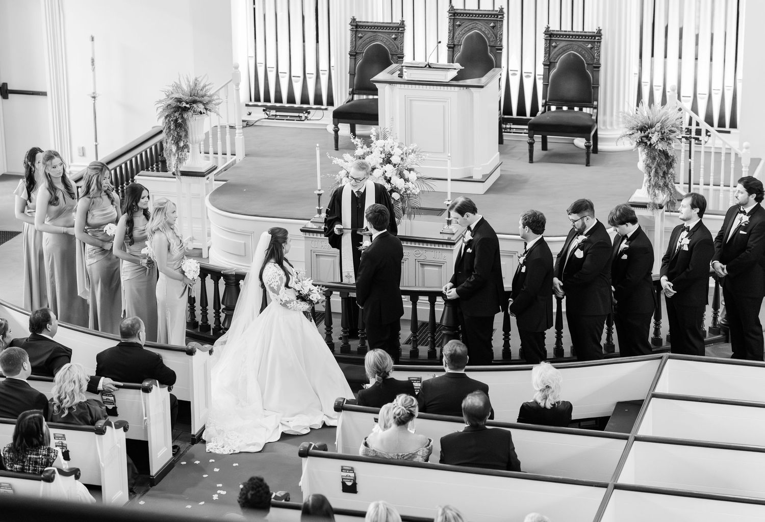 Black and white photo of bride and groom exchanging vows at church altar with wedding party during ceremony.
