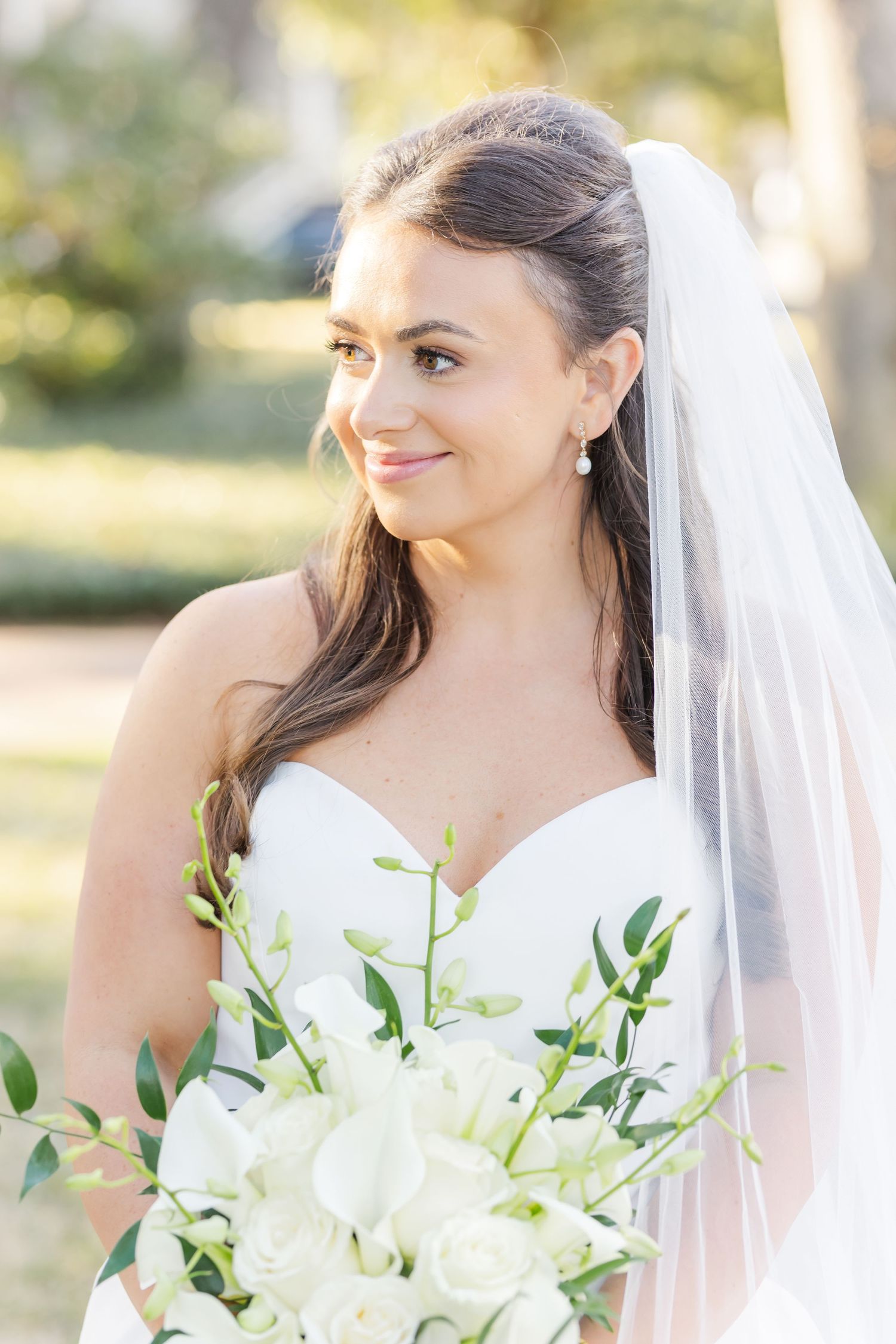 Beautiful bride with dark hair, veil, and white floral bouquet smiling softly in outdoor setting.