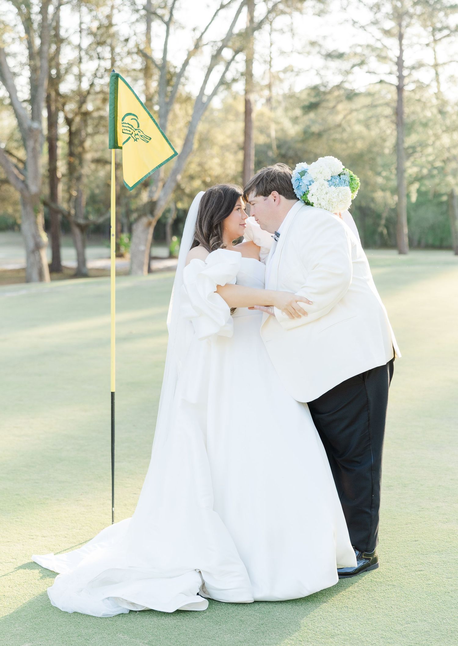 Bride and groom share a romantic kiss on a golf course green beside a yellow flag on their wedding day.