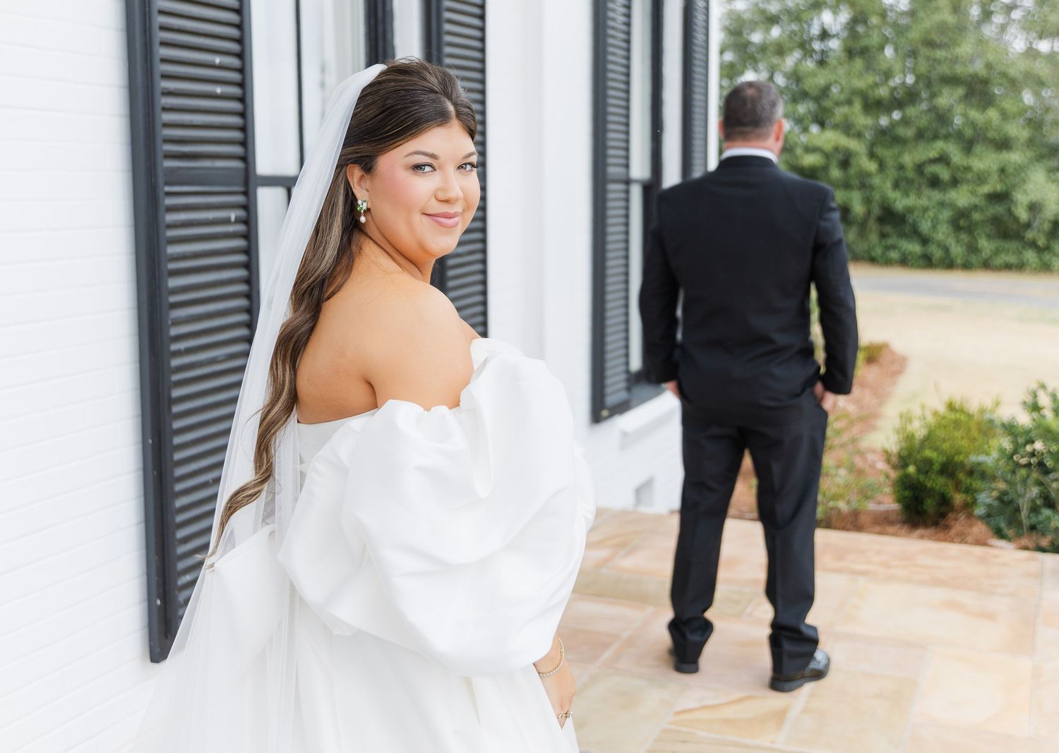 Bride in off-shoulder white gown smiles over shoulder during first look moment at elegant wedding venue.