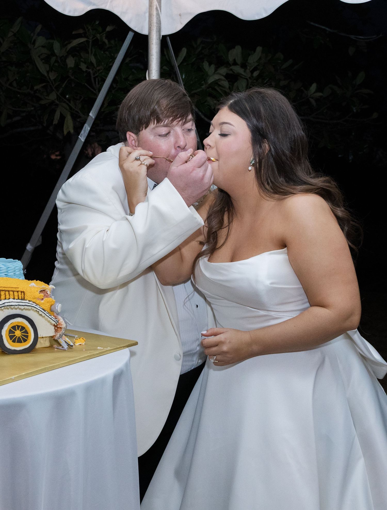Bride and groom share a joyful cake cutting moment at their outdoor wedding reception under a white tent.