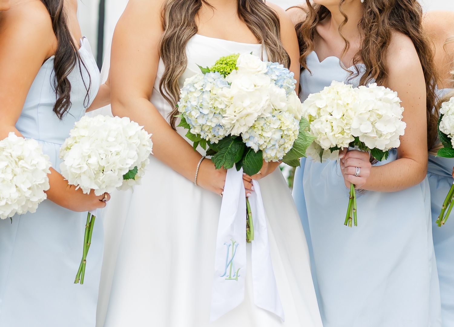 Bride and bridesmaids in light blue dresses hold white hydrangea bouquets, bride displaying monogrammed ribbon detail.