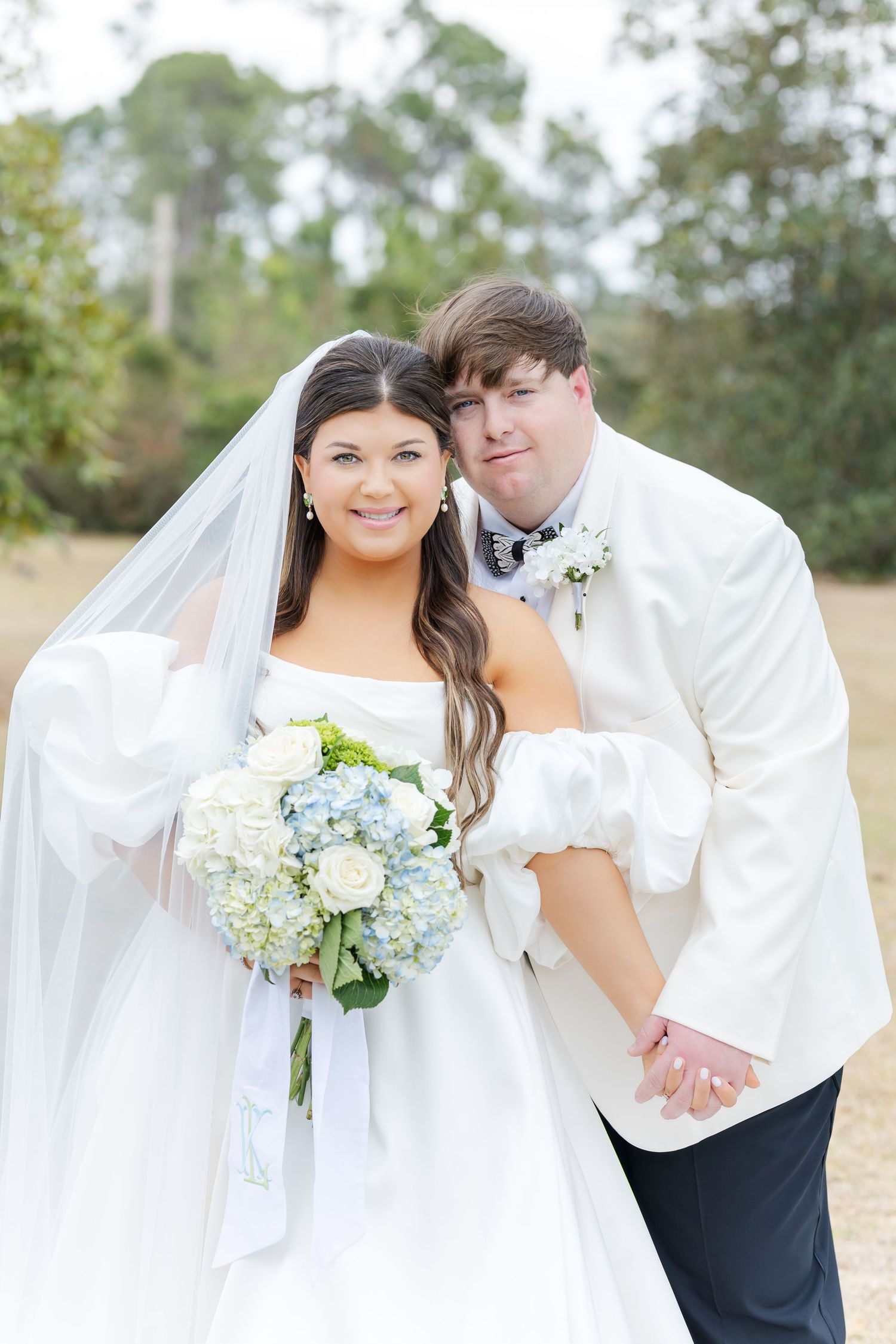 Smiling bride in off-shoulder wedding gown holding blue and white hydrangea bouquet posing with groom in ivory tuxedo jacket.