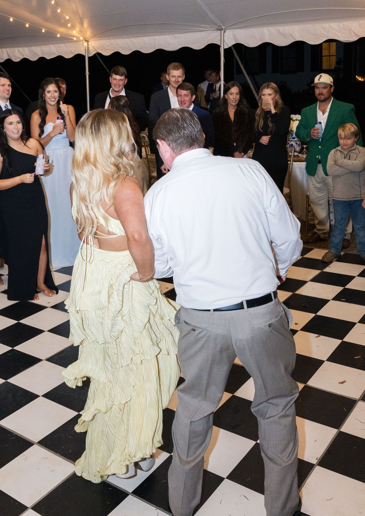 Couple dances on black and white checkered floor at outdoor wedding reception as guests watch under white tent.