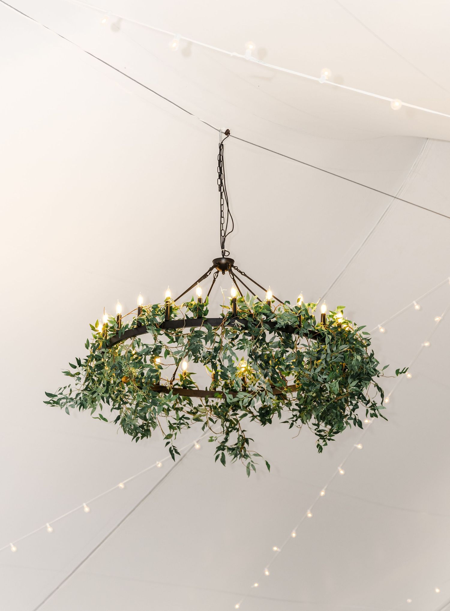 Rustic chandelier adorned with lush greenery and candles hanging from ceiling with string lights at wedding venue.