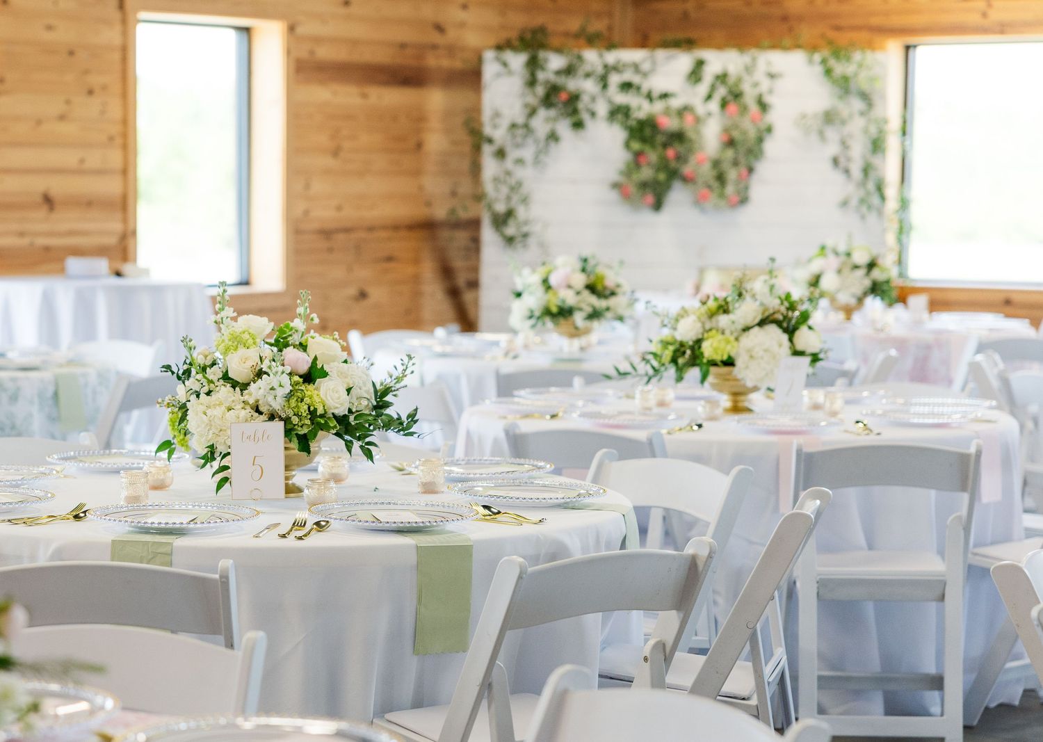 Elegant wedding reception hall with white floral centerpieces, green table runners, and white chairs in a rustic barn venue.