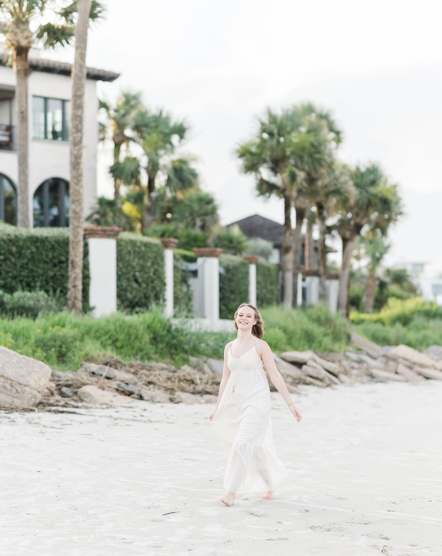 A person in a white dress walks along a sandy beach with palm trees and coastal homes in the background.