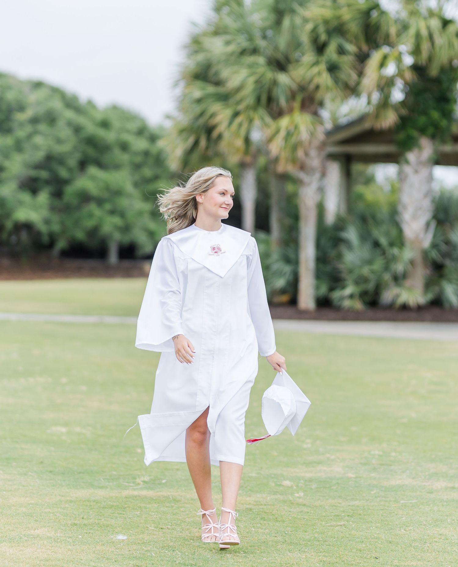 Graduate in white cap and gown walking through a grassy field with palm trees in the background.