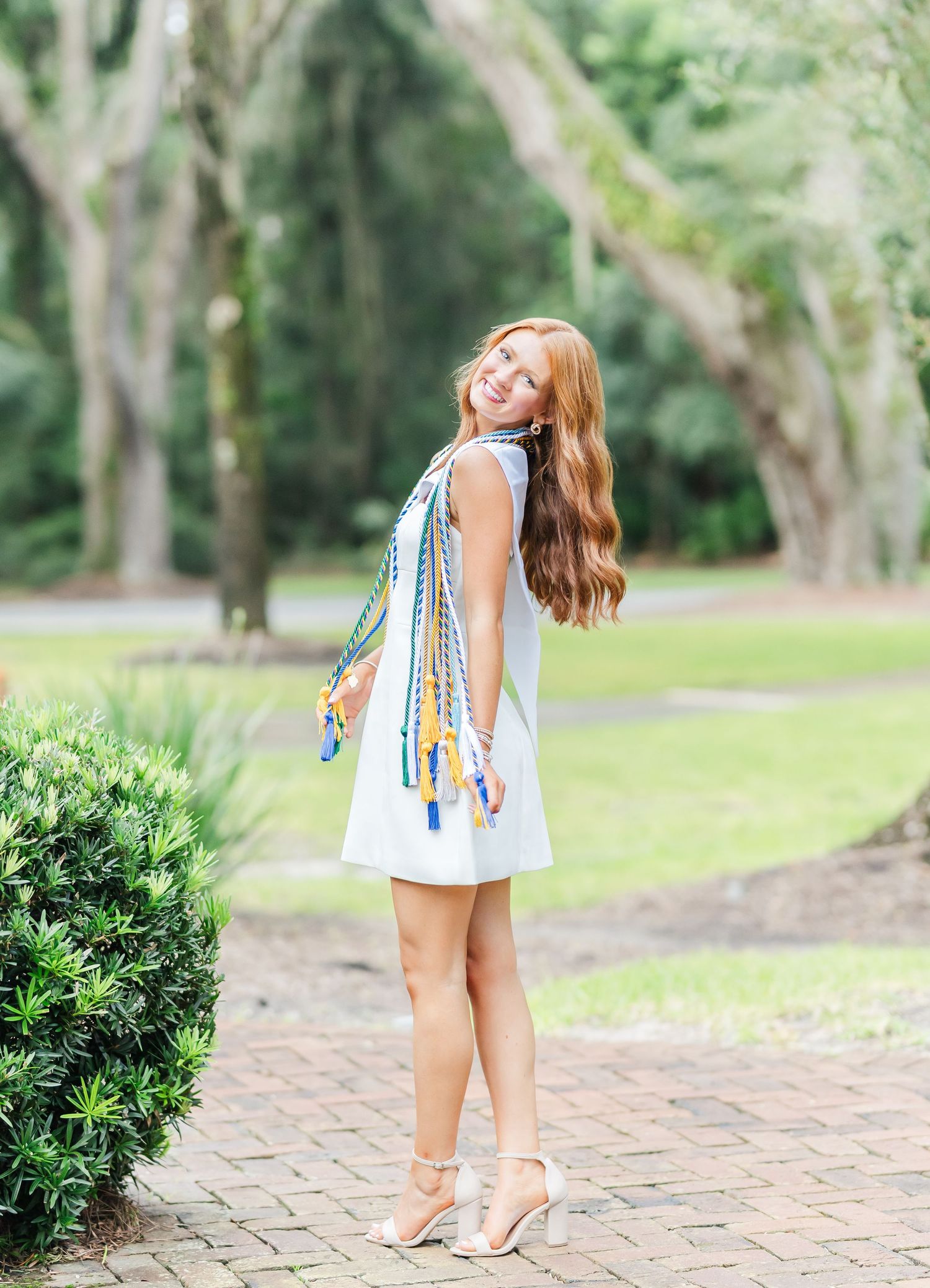 Girl in white dress with colorful braided details stands on garden pathway near greenery.