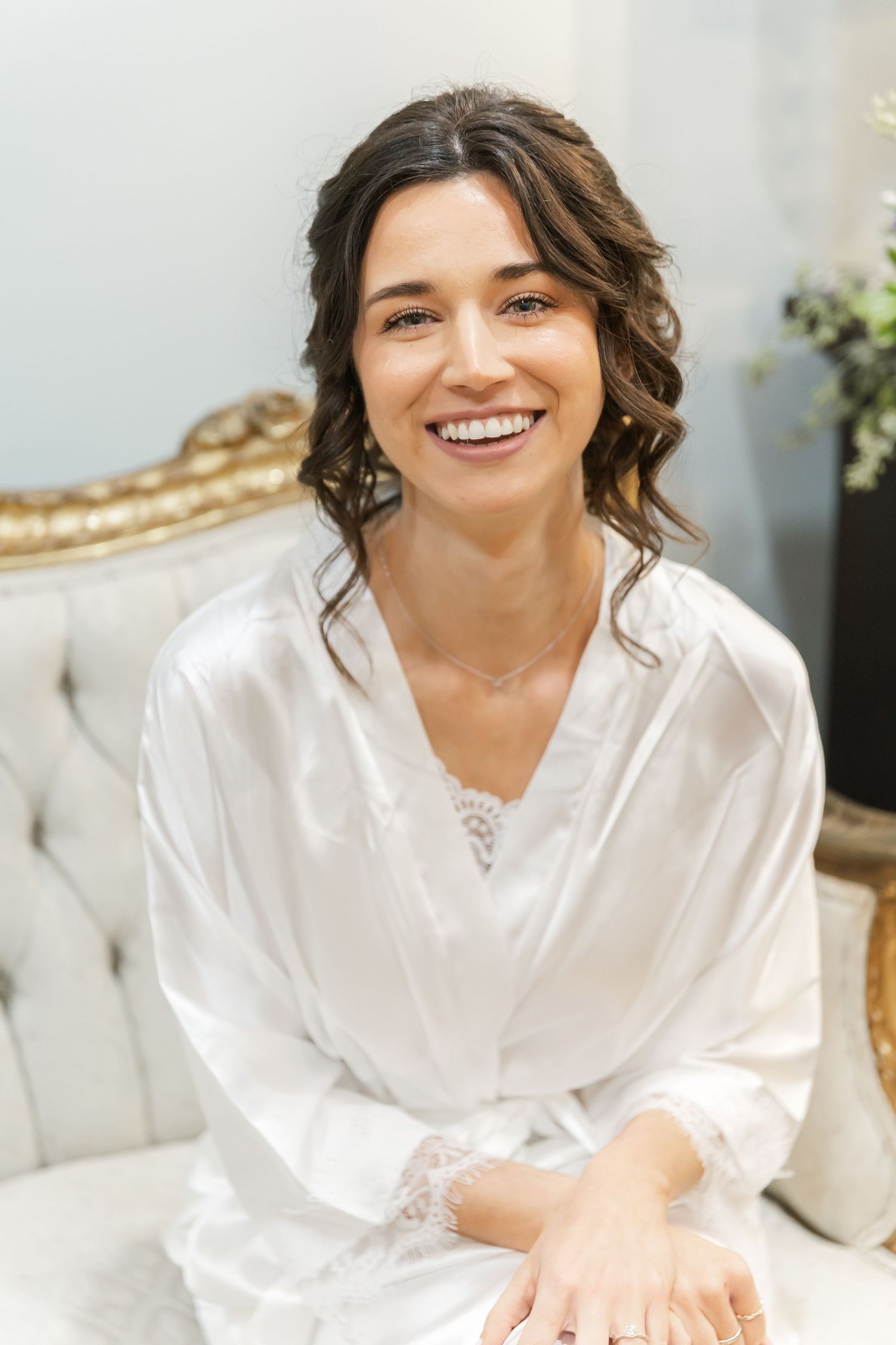A person wearing a white silk robe sits on a couch smiling warmly in a bright indoor setting.