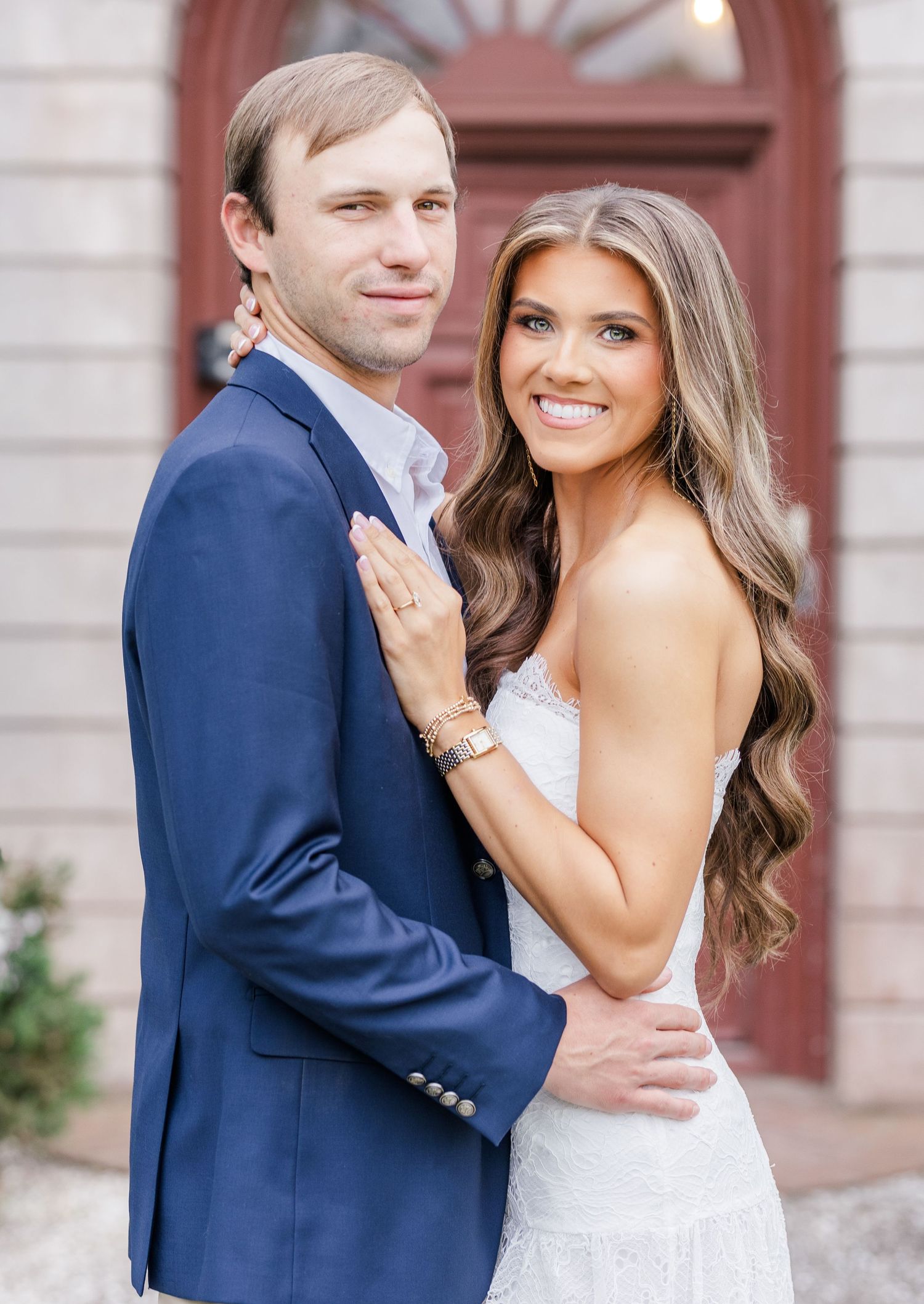 Smiling couple in engagement photo, man in navy suit, woman in white lace dress with long wavy hair.