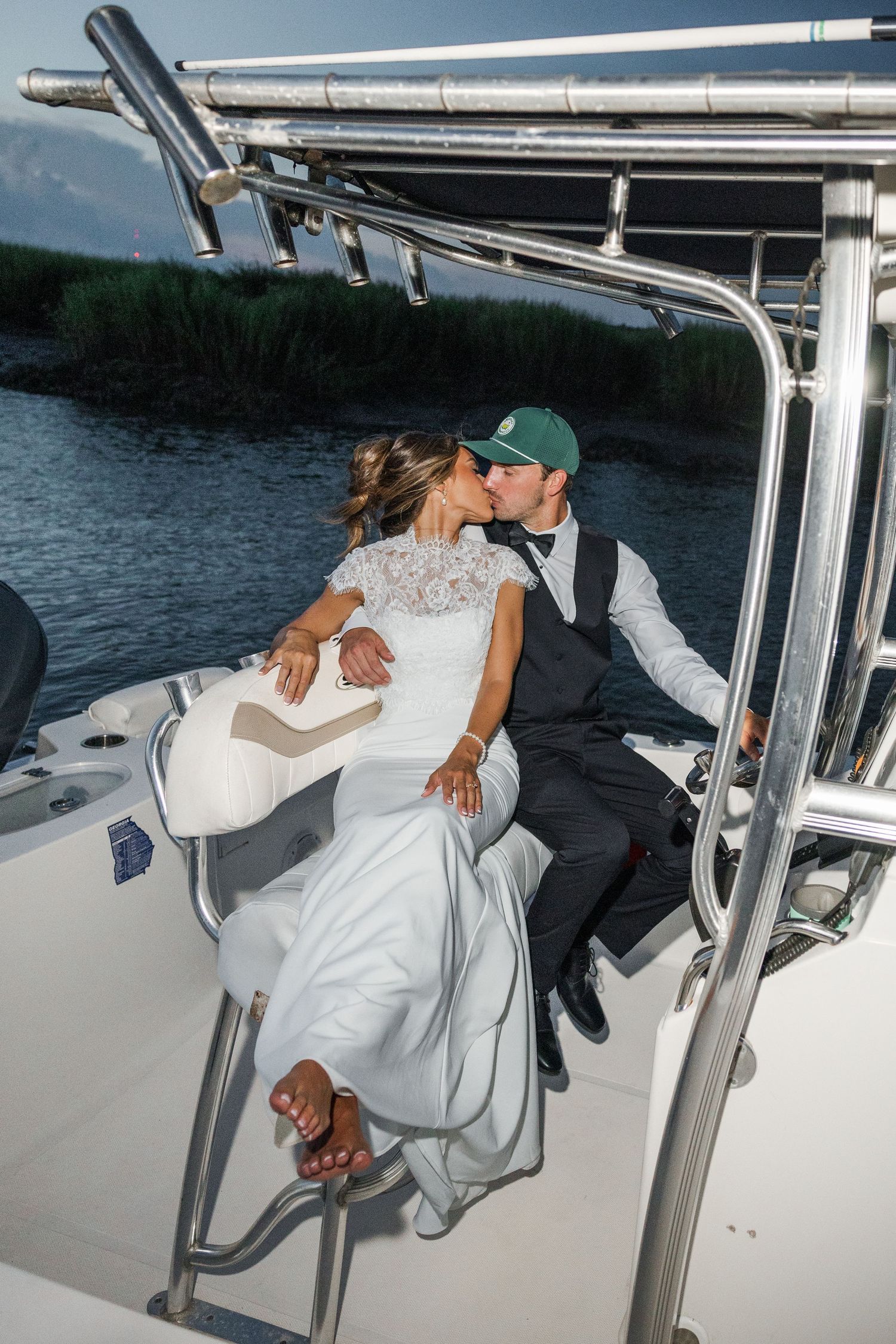 Bride and groom sharing a kiss on a white boat near a marsh waterway, wearing wedding attire.