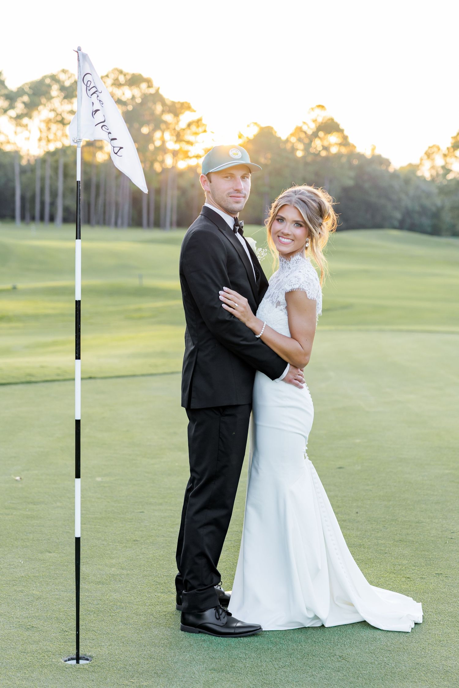 Bride and groom posing on a golf course green at sunset, she in a mermaid gown, he in a black tuxedo.