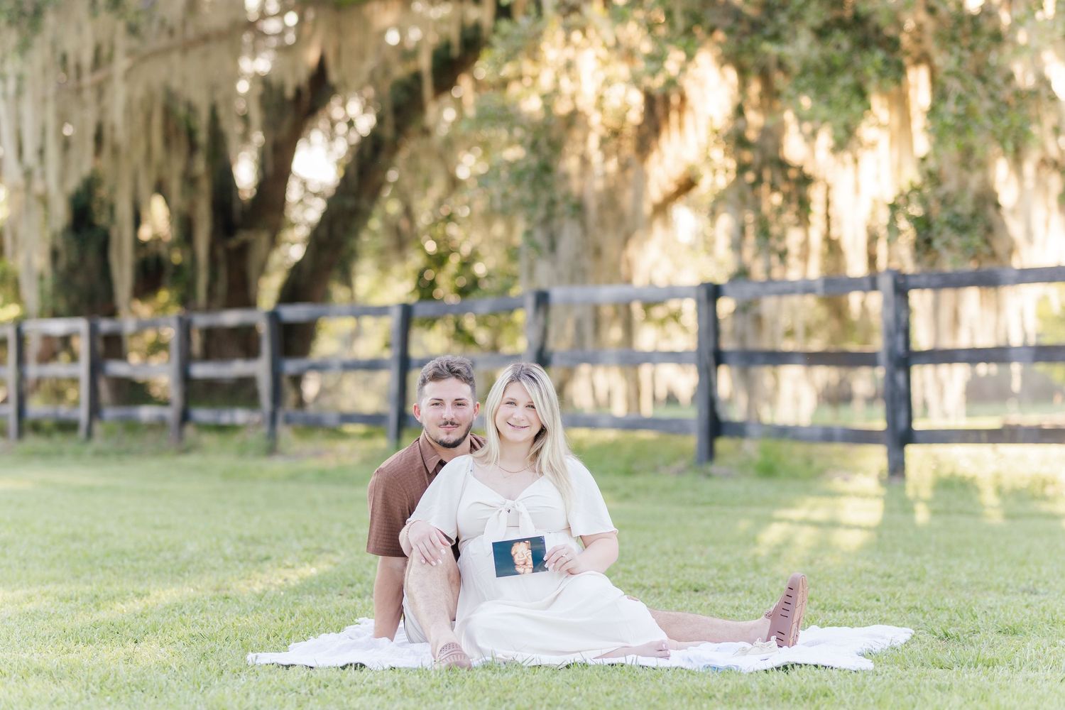 Couple sitting on white blanket in grassy field with Spanish moss oak tree and wooden fence backdrop.