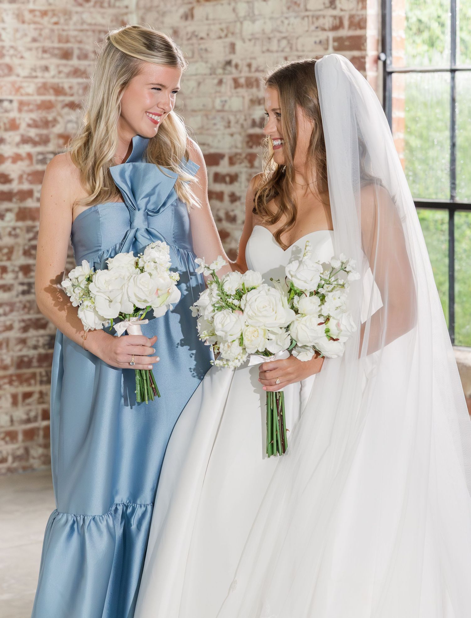 Bride in white gown with veil and bridesmaid in blue bow dress holding white bouquets indoors.