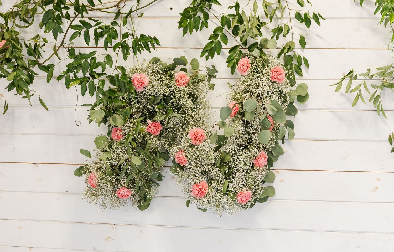 Floral letter M decoration made with pink roses and greenery mounted on a white wooden wall.
