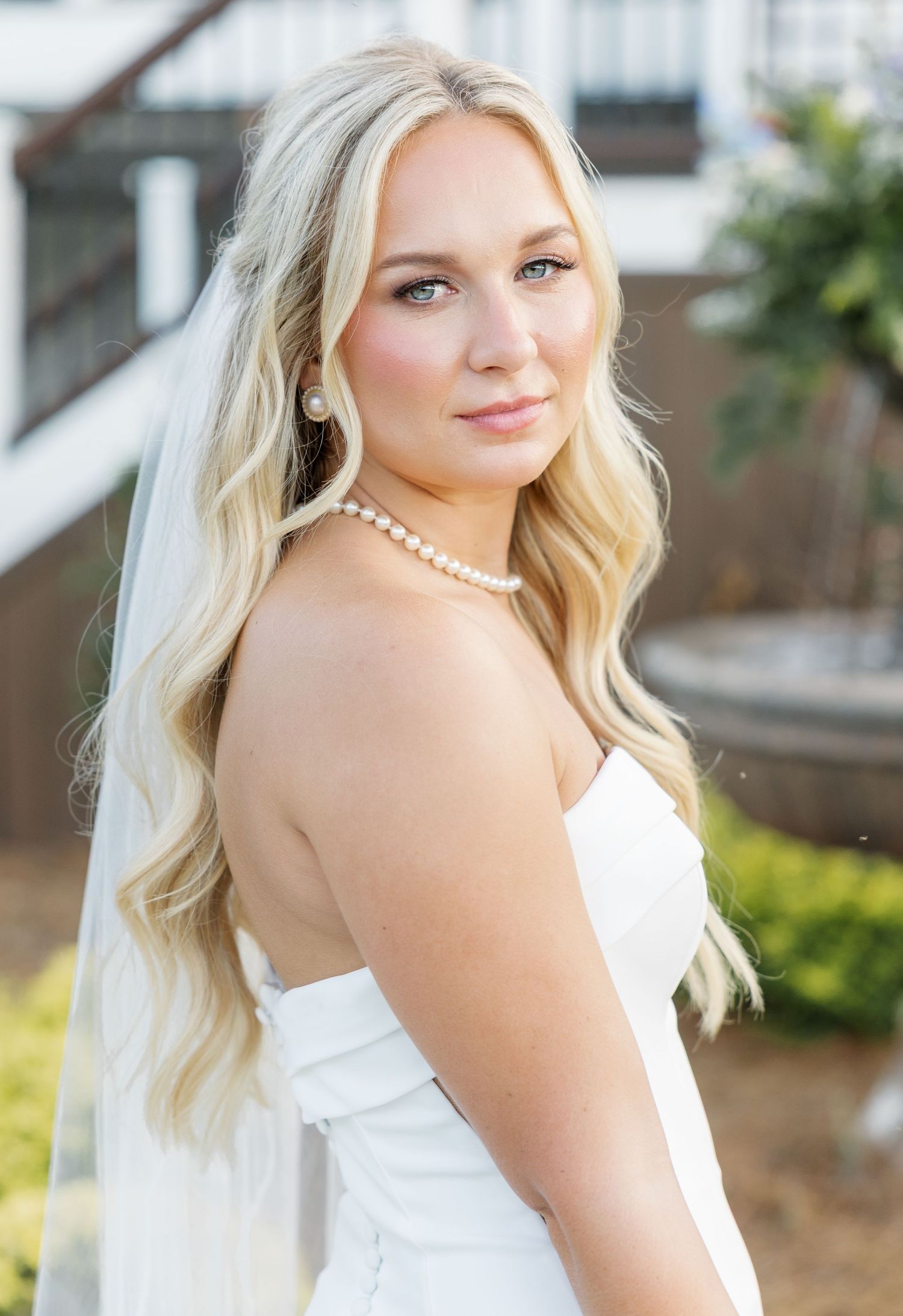 Stunning blonde bride in strapless gown with pearl necklace and veil posing outdoors at wedding venue.