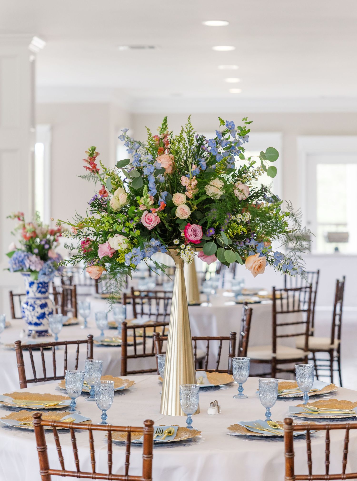 Tall gold vase with lush pink, peach, and blue floral centerpiece on white-clothed reception table.