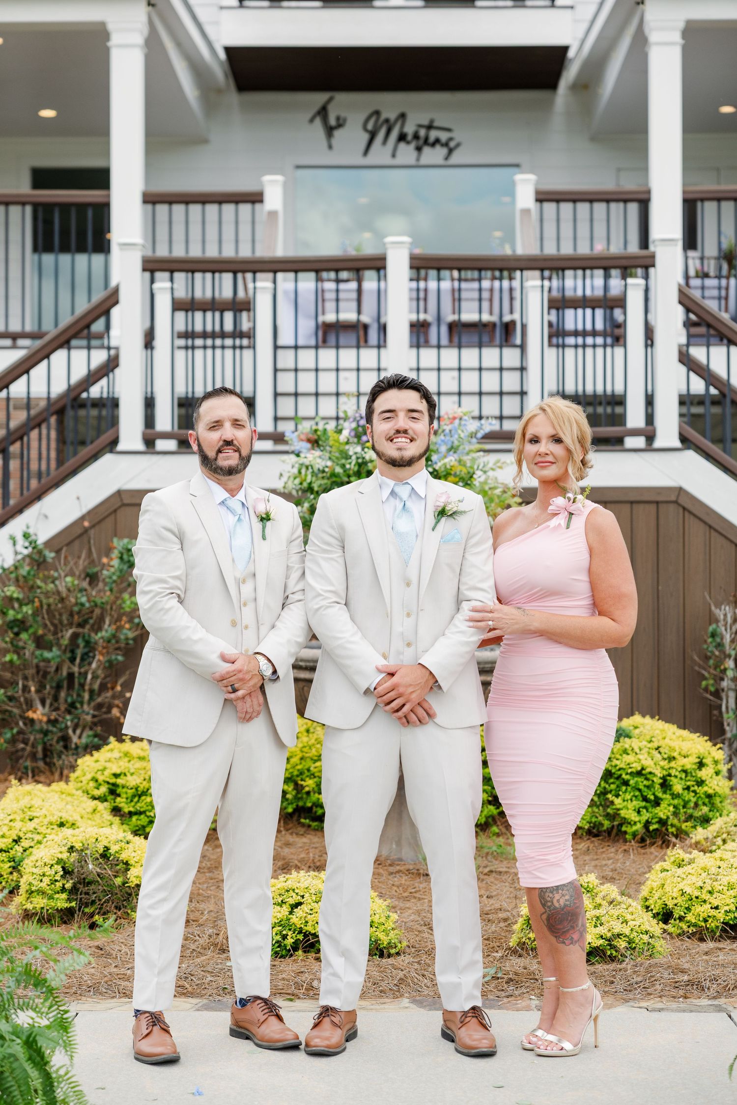 Groom in white suit poses with groomsman and woman in pink dress outside wedding venue entrance.