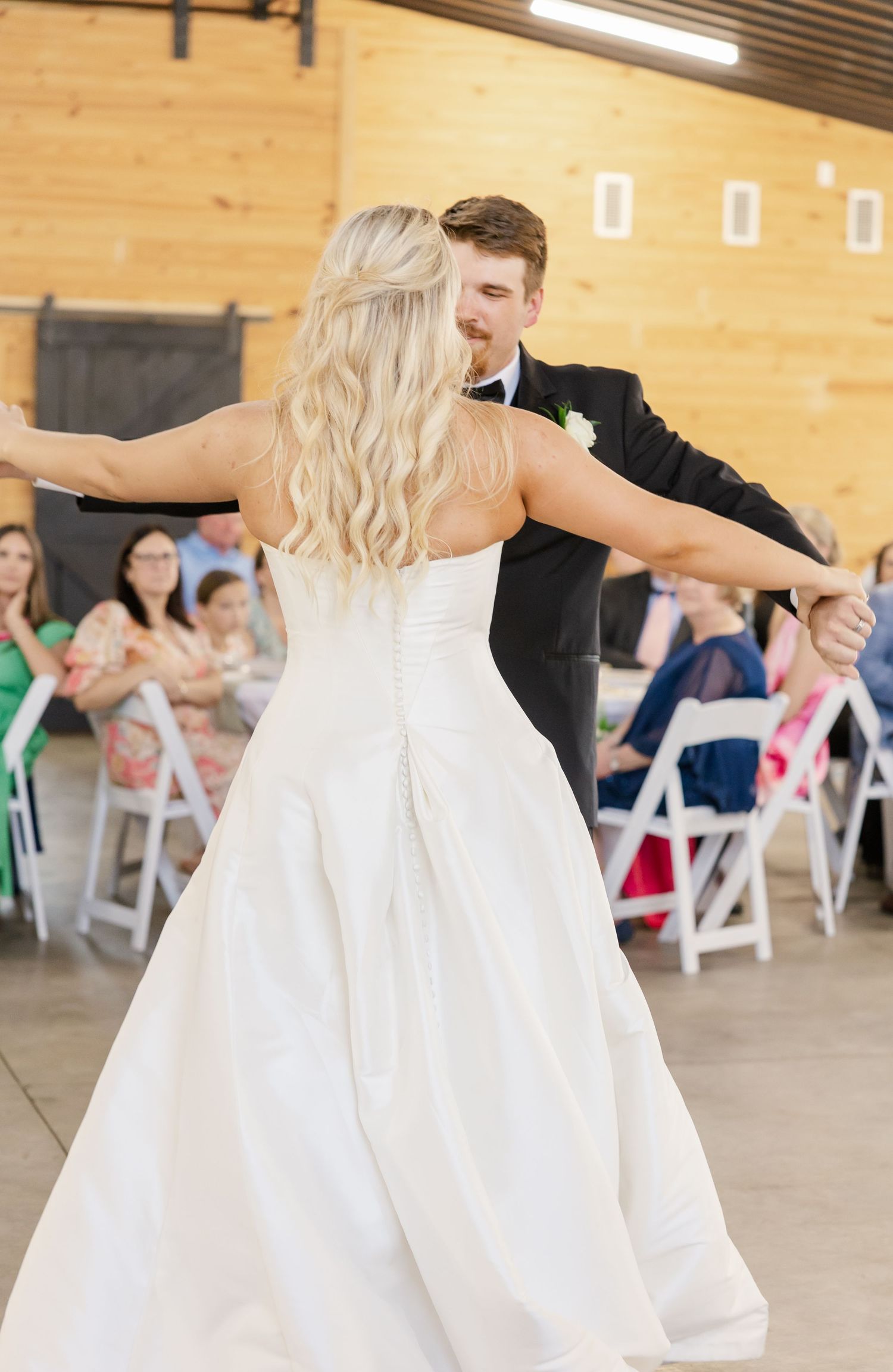 Bride in strapless white ball gown dancing with groom in black tuxedo at barn reception.