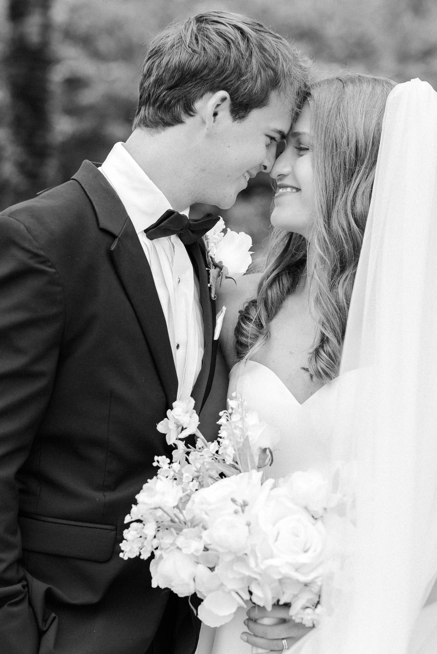 Black and white photo of bride and groom sharing an intimate moment, touching foreheads while bride holds white floral bouquet.