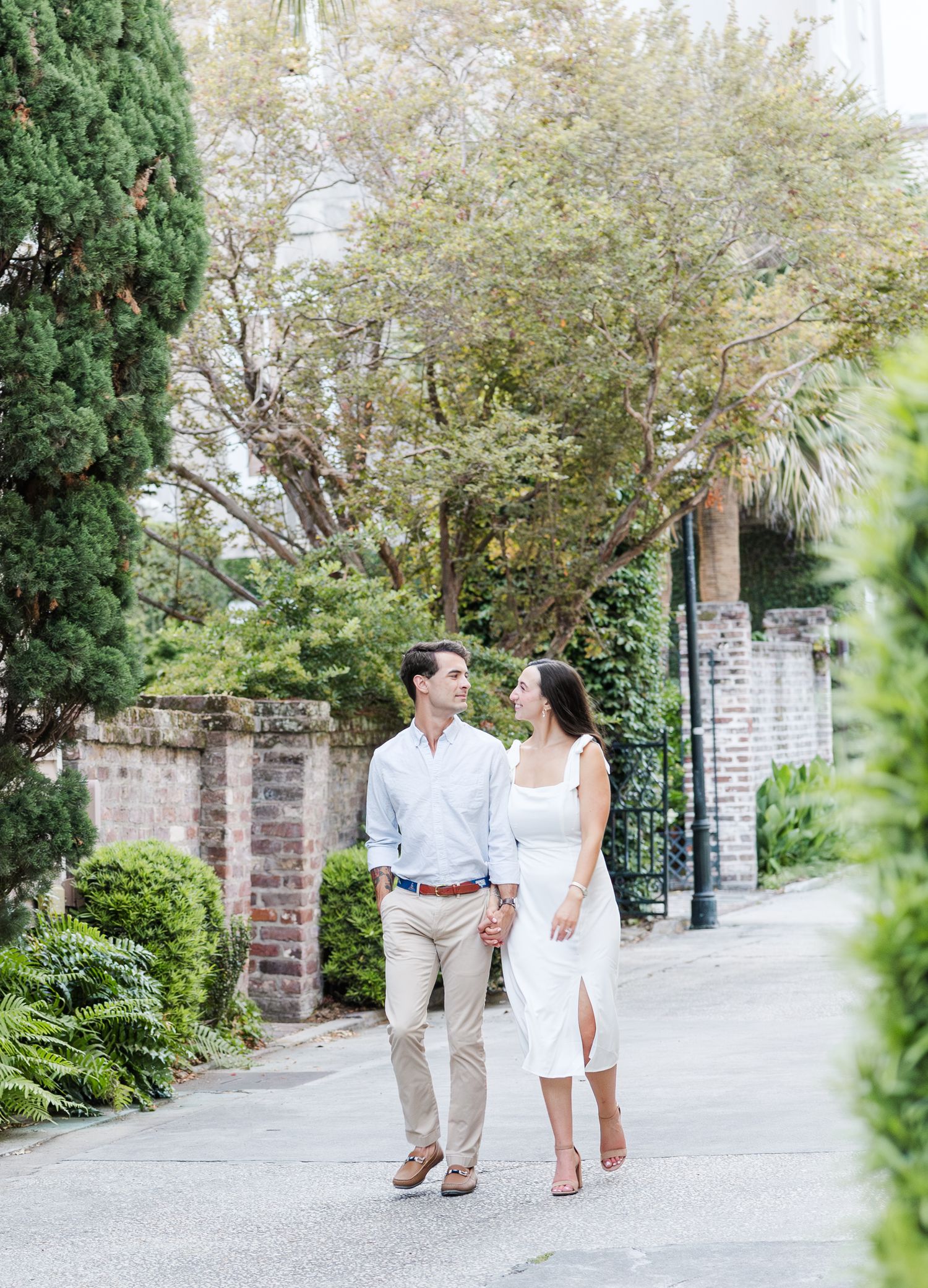 Couple holding hands and gazing at each other while strolling along a lush tree-lined brick pathway.