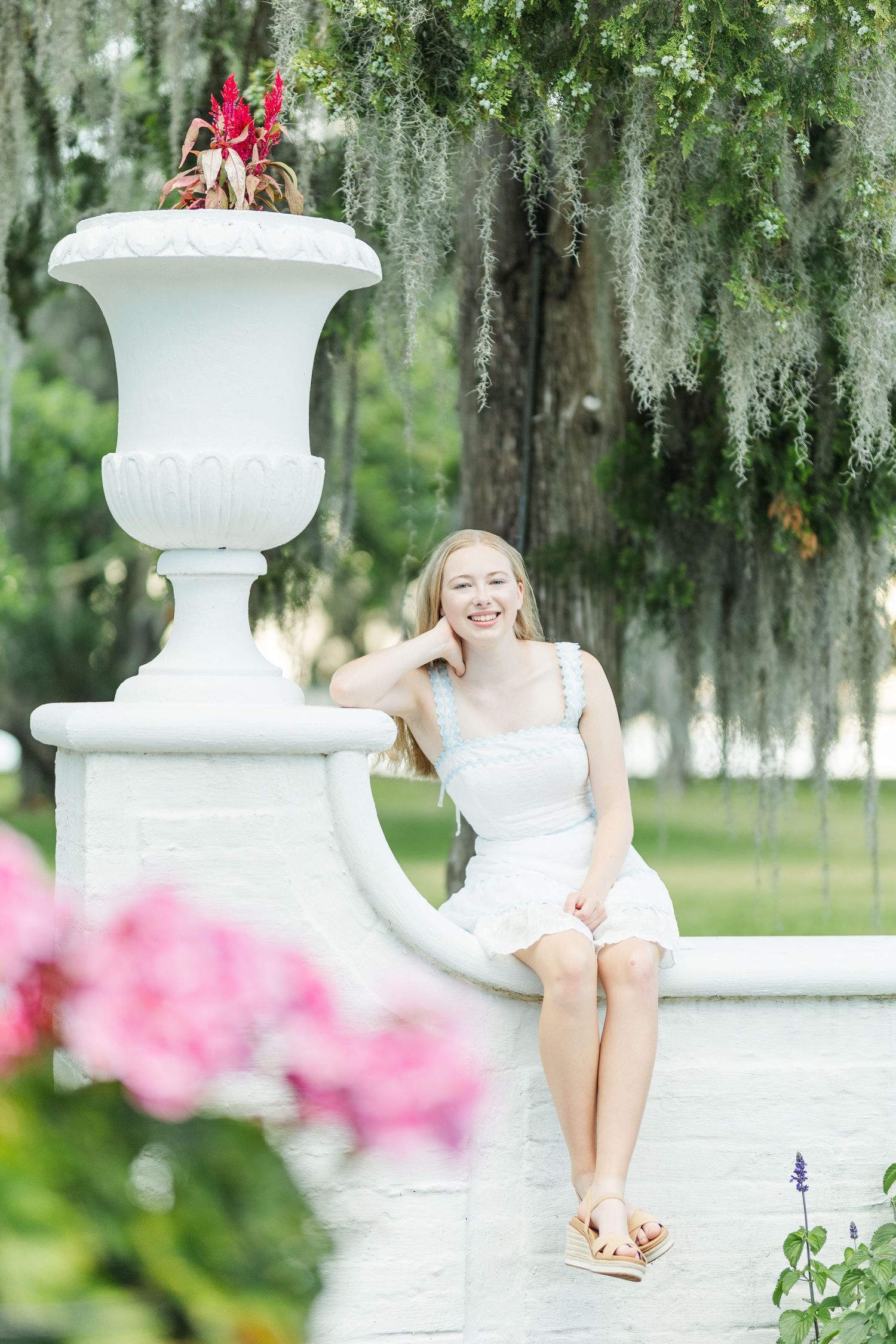 Three white decorative columns in a garden with pink flowers and Spanish moss hanging from trees in the background.